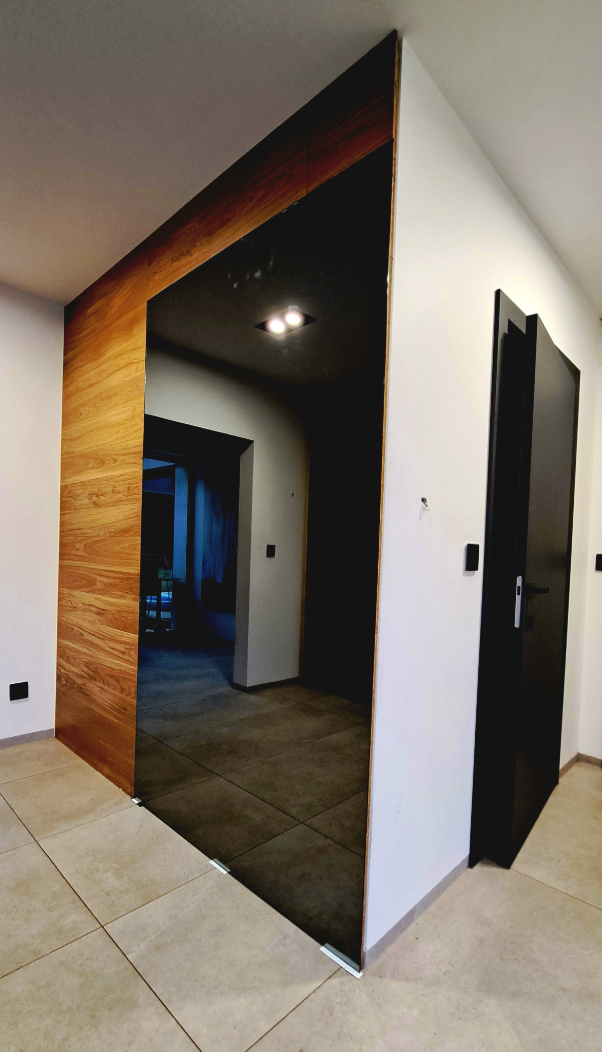 Nowoczesne wnętrze z przesuwnymi drzwiami szklanymi z drewnianą ramą, odbijającymi dalszą część korytarza z widoczną czarną ścianą i drzwiami.