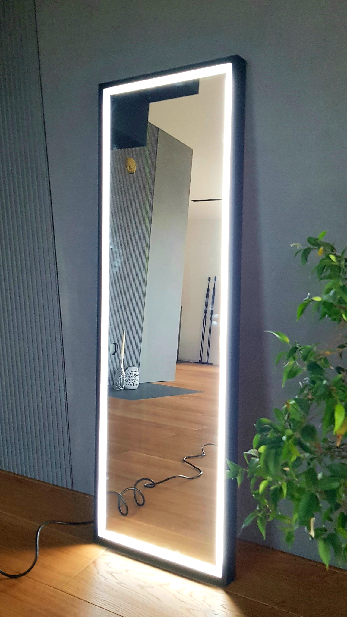 Wysokie, prostokątne lustro podświetlane diodami LED, oparte o szarą ścianę i drewnianą podłogę, z widocznym kablem zasilającym i odbiciem fragmentu pokoju.