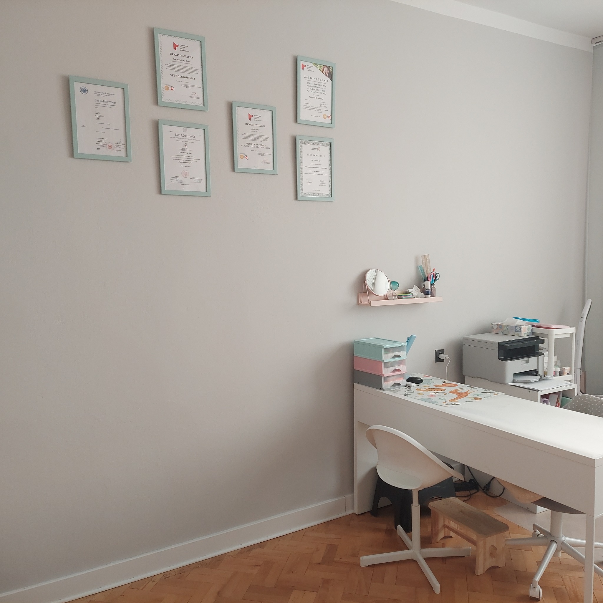 Gabinet z biurkiem, krzesłami, półką z akcesoriami i ścianą z oprawionymi dyplomami i certyfikatami, prawdopodobnie gabinet specjalisty.