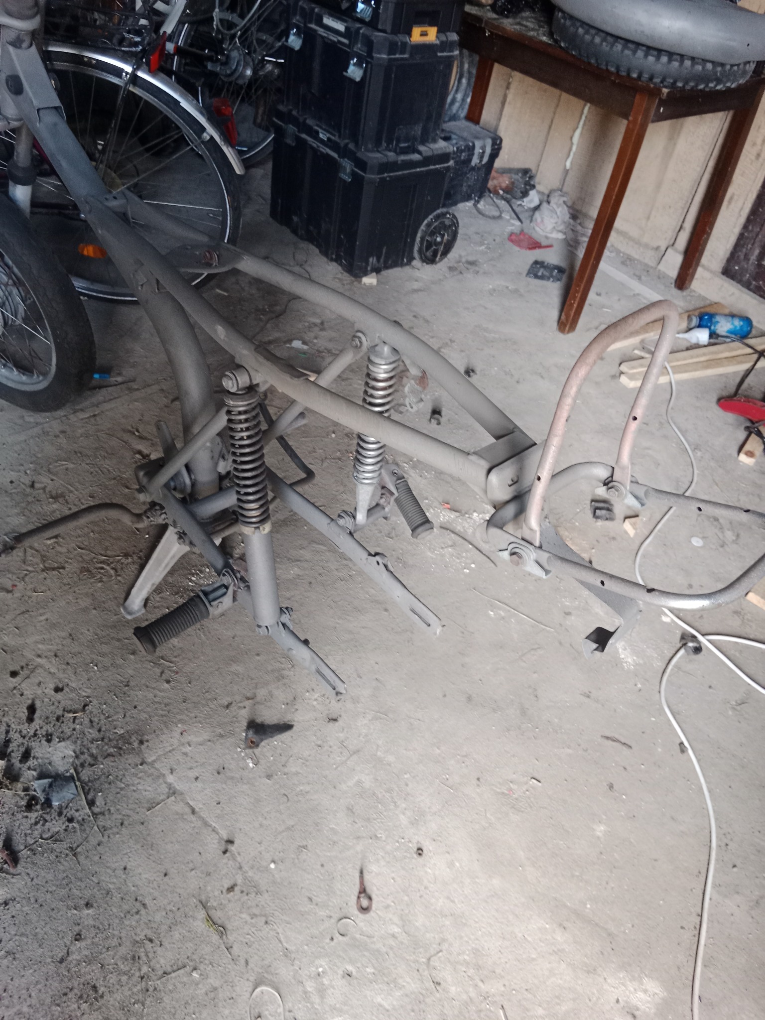 Rama wózka inwalidzkiego lub trójkołowca pokryta szarym podkładem, widoczne sprężyny zawieszenia, w tle rowery i narzędzia warsztatowe.