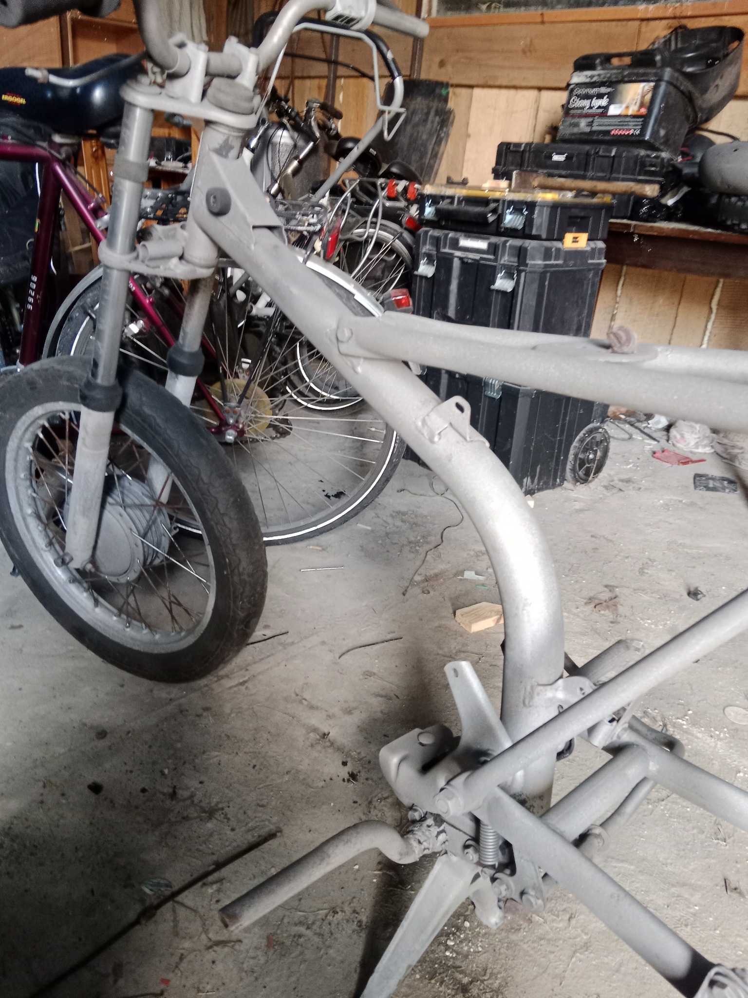 Rama motocykla po piaskowaniu, przygotowana do malowania proszkowego; widoczne ślady po piaskowaniu i szary matowy odcień, w tle inne rowery i skrzynki narzędziowe.