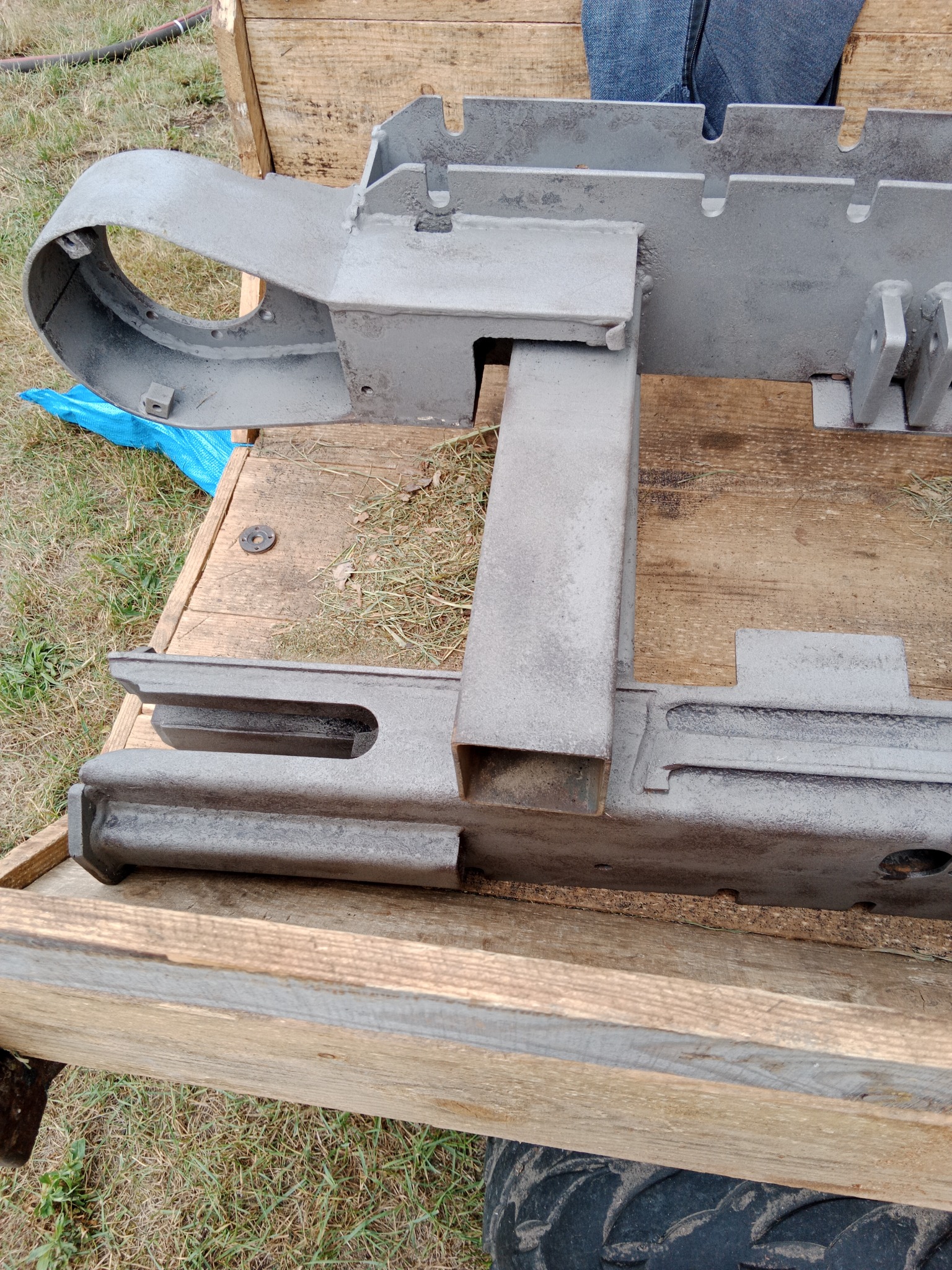 Szary, metalowy element konstrukcyjny, prawdopodobnie fragment maszyny, leżący na drewnianej skrzyni, po obróbce ściernej.