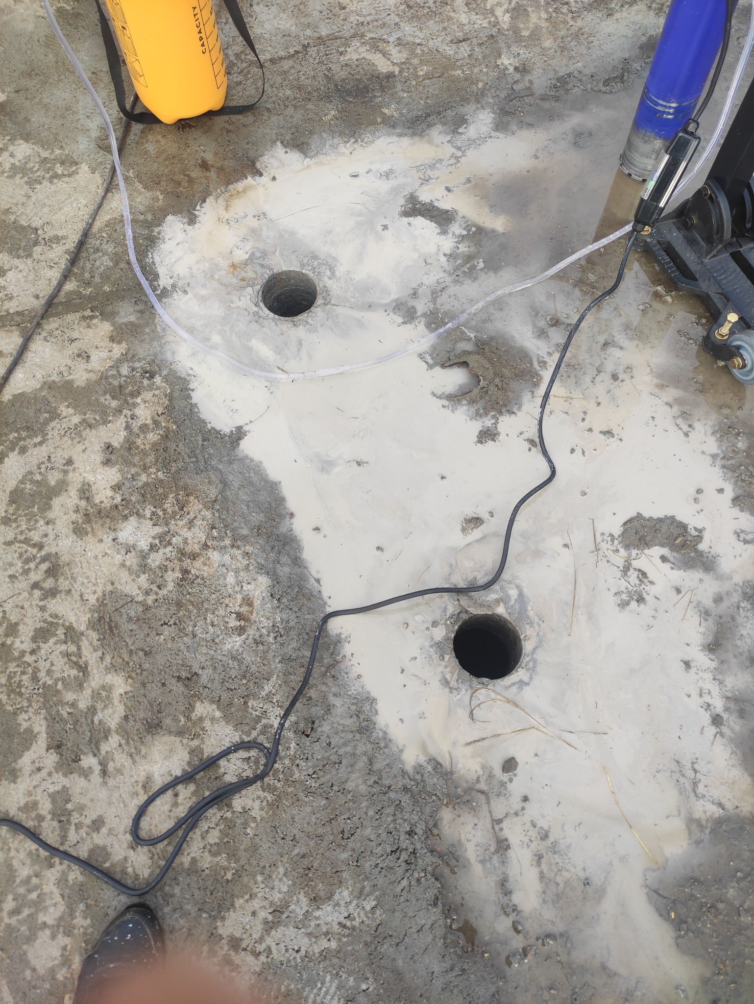 Dwa wywiercone otwory w betonowej powierzchni, otoczone mieszaniną wody i pyłu, z widocznym sprzętem w tle i żółtym pojemnikiem na górze.