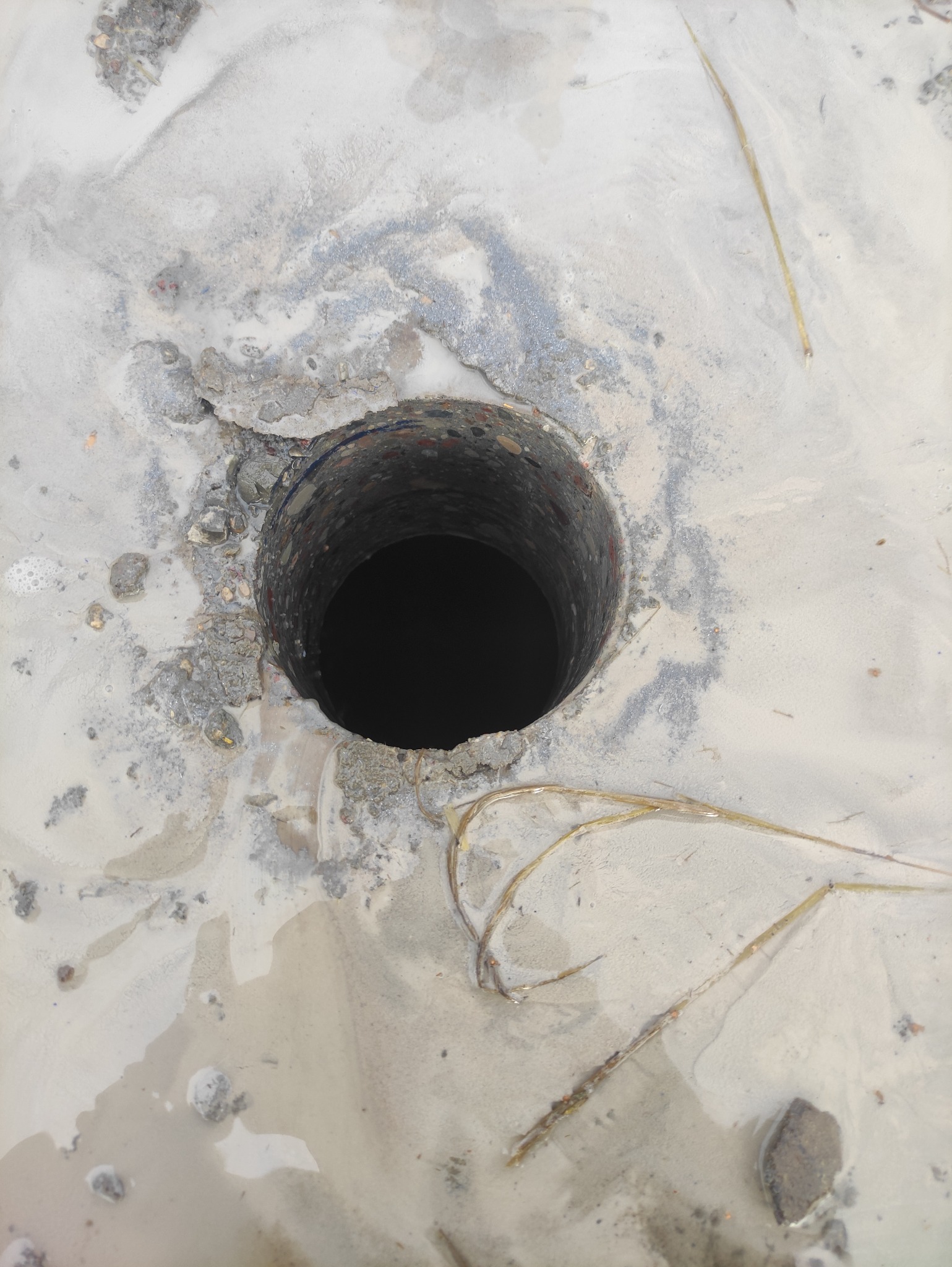 Okrągły wykop w ziemi z widocznymi śladami wody i fragmentami gruzu wokół krawędzi. Ujęcie z góry, podkreślające głębię otworu i surowość otoczenia.