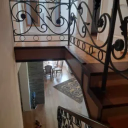 Kuta, czarna balustrada z motywem kwiatowym na schodach, widok z góry na drewniane stopnie i fragment jadalni z dywanem w tle.