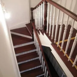 Klatka schodowa z ciemnego drewna z białymi listwami przypodłogowymi i białą balustradą z brązowym poręczą, widok z góry na zakręcające schody.