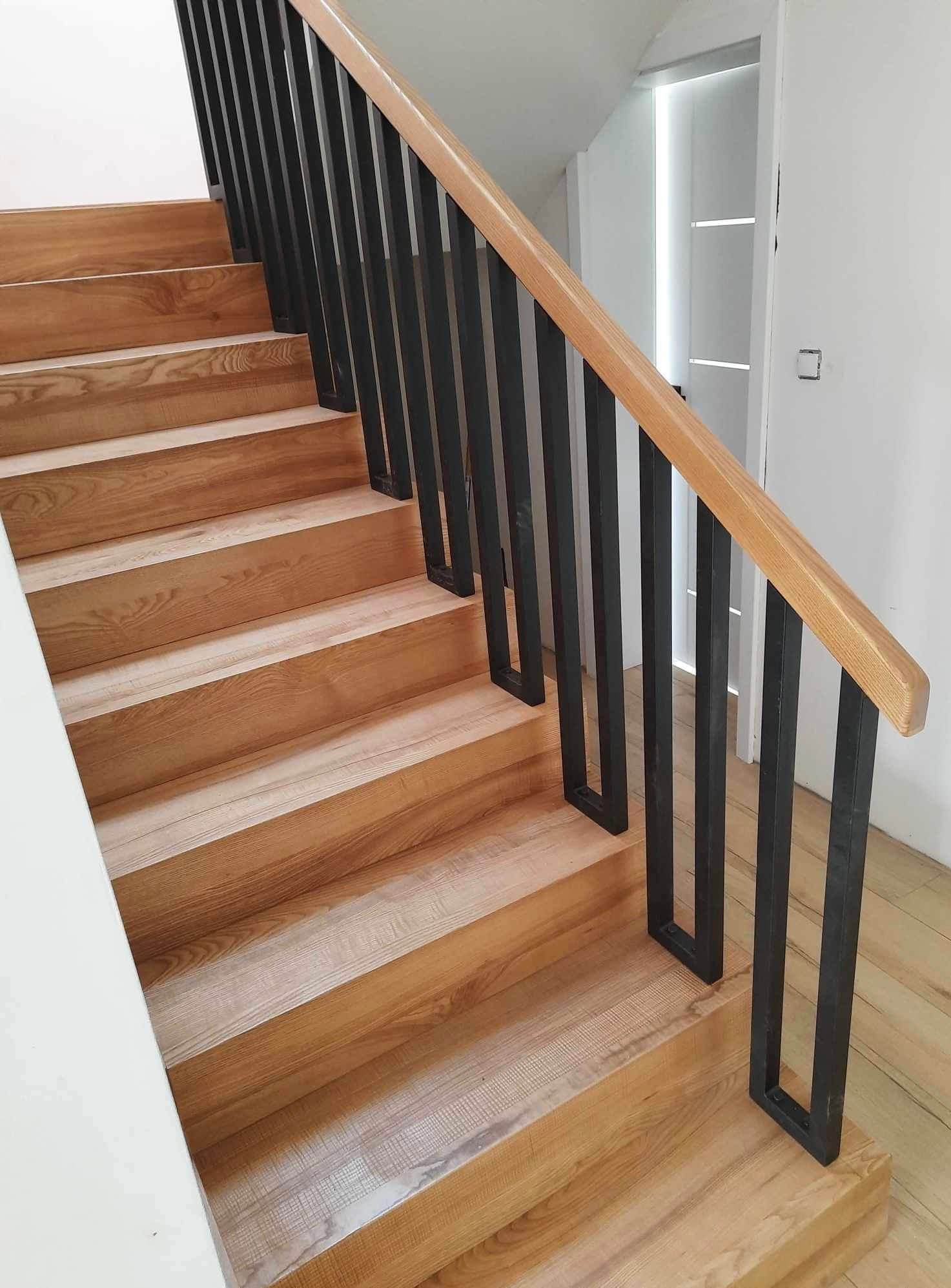 Drewniane schody z czarnymi, metalowymi balustradami i jasną, drewnianą poręczą. Widoczne jasne stopnie i podłoga, białe ściany i drzwi w tle.