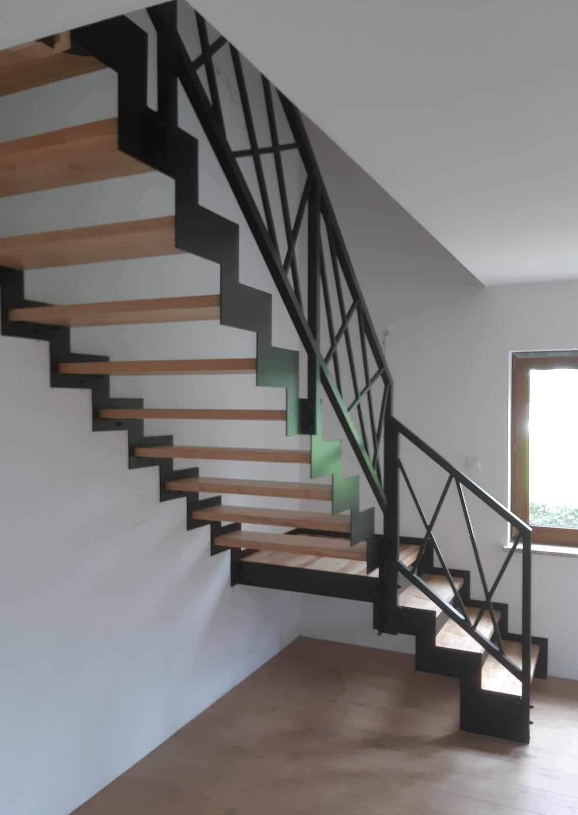 Nowoczesne schody z drewnianymi stopniami i czarną, metalową konstrukcją oraz balustradą o geometrycznym wzorze, umieszczone w jasnym wnętrzu z oknem.