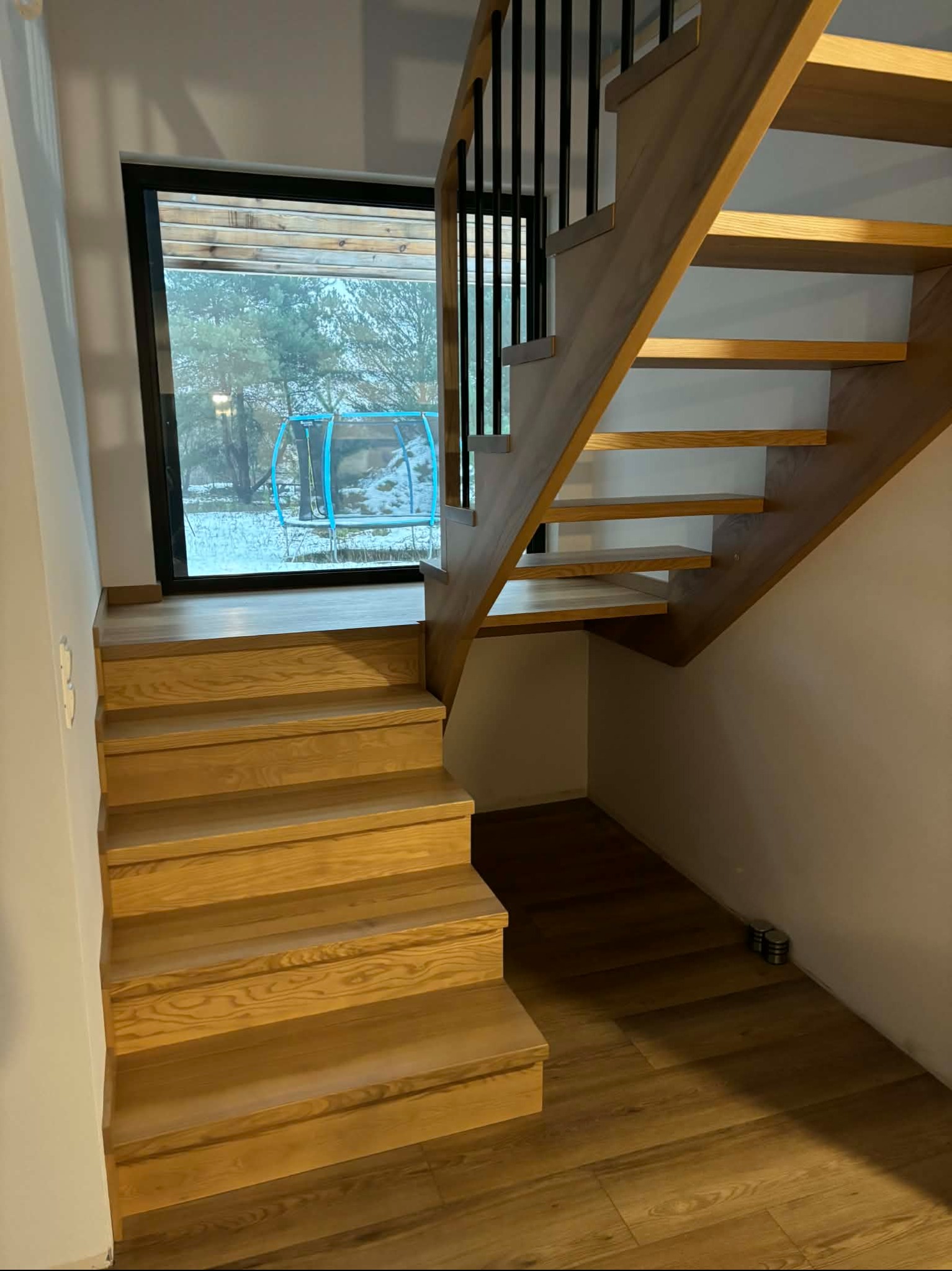 Drewniane schody z czarną balustradą w domu w Rąbiniu, widoczne przez okno drzewa i trampolina. Naturalne wykończenie drewna, nowoczesny design.