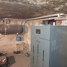 Eko Serwis - Szary piec Ferroli w piwnicy z instalacją CO, zbiornikiem Kospel i rurami. Widoczne narzędzia i akcesoria instalacyjne. Surowy betonowy sufit i ściany.