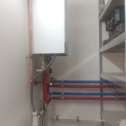 Eko Serwis - Biały piec gazowy Viessmann z miedzianymi i plastikowymi rurami (czerwone, niebieskie) zamontowany na ścianie w pomieszczeniu gospodarczym, obok metalowy regał.