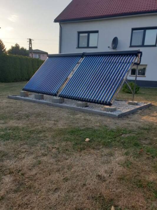 Dwa kolektory słoneczne rurowe zamontowane na trawniku przed domem z czerwoną dachówką. Widoczne betonowe podstawy i metalowe wsporniki.