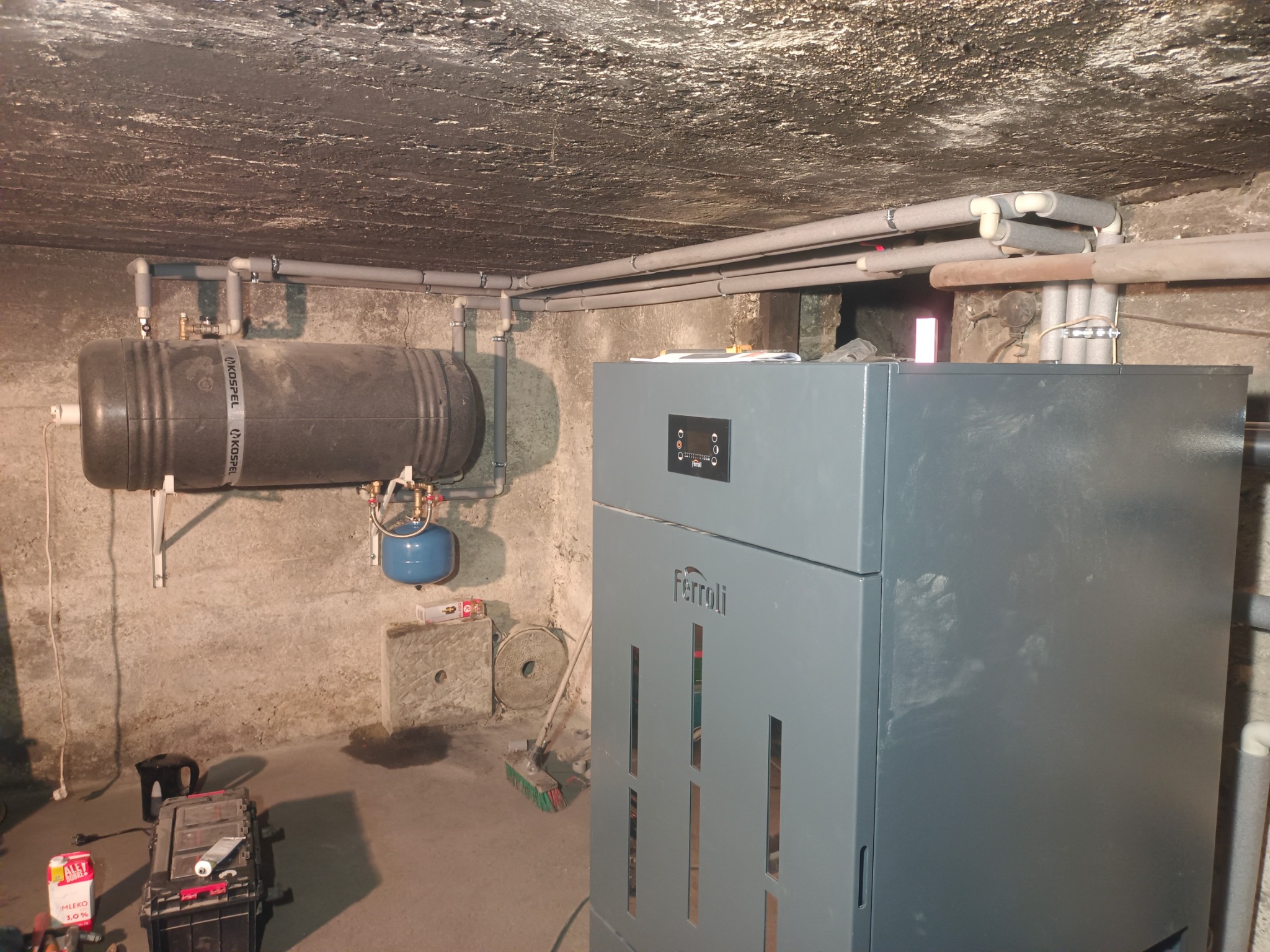 Szary piec Ferroli w piwnicy z instalacją CO, zbiornikiem Kospel i rurami. Widoczne narzędzia i akcesoria instalacyjne. Surowy betonowy sufit i ściany.