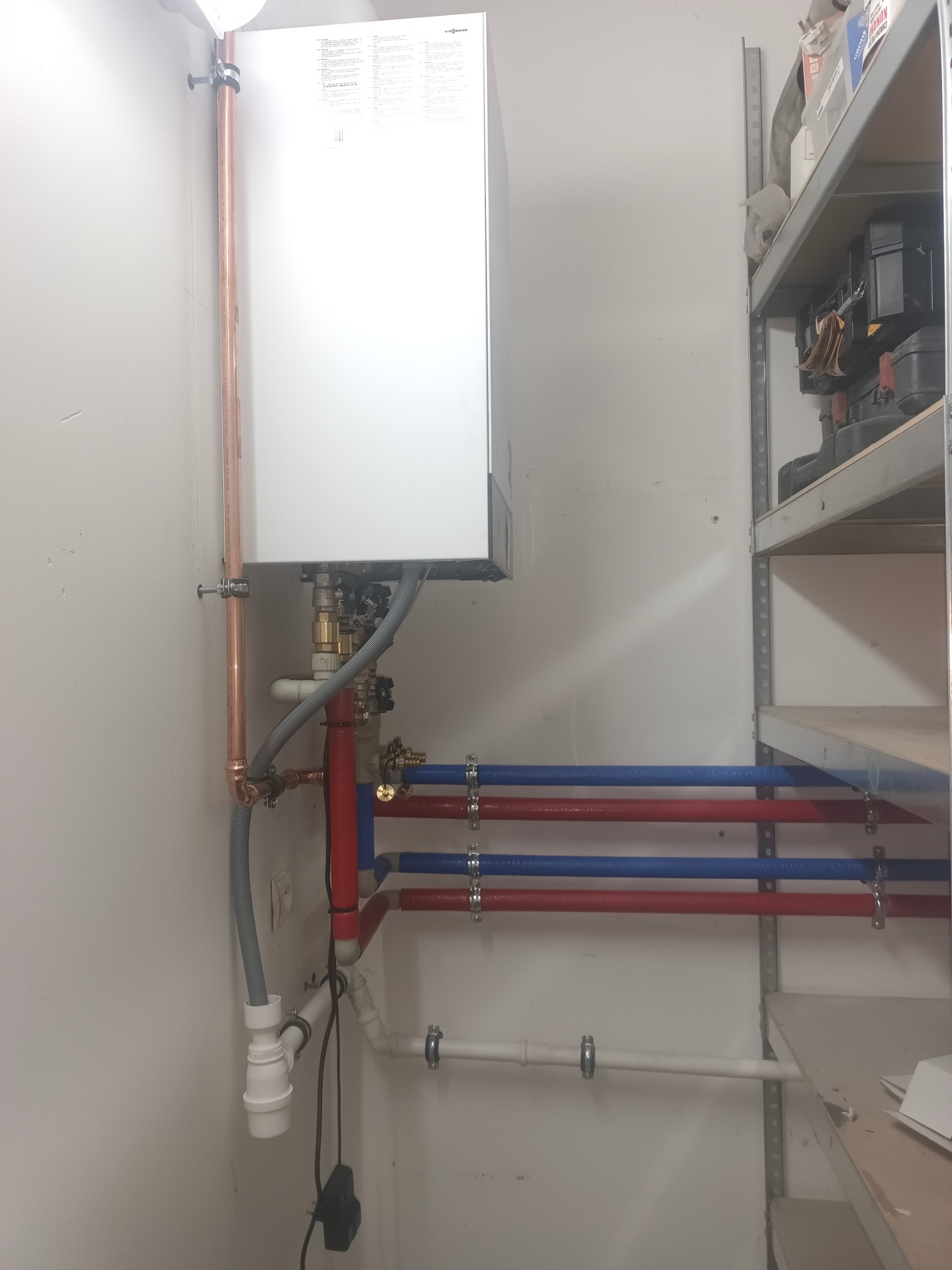 Biały piec gazowy Viessmann z miedzianymi i plastikowymi rurami (czerwone, niebieskie) zamontowany na ścianie w pomieszczeniu gospodarczym, obok metalowy regał.