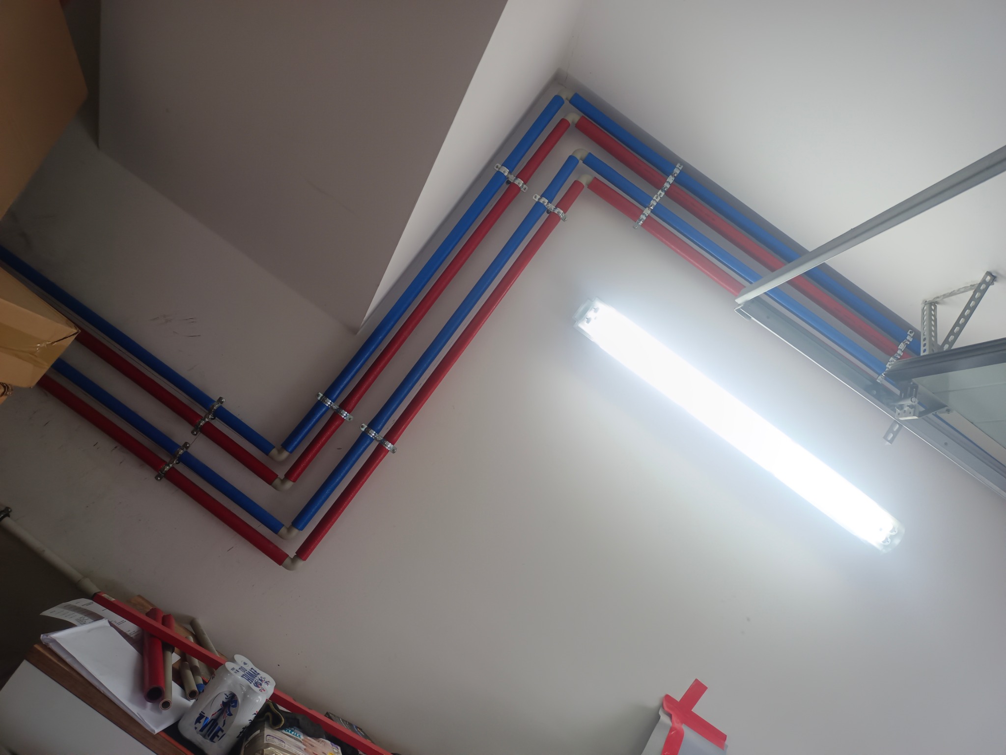 Instalacja rur PEX (czerwone i niebieskie) pod sufitem w Kętach, zabezpieczone metalowymi uchwytami. Widoczna lampa oświetleniowa i fragment konstrukcji bramy garażowej.