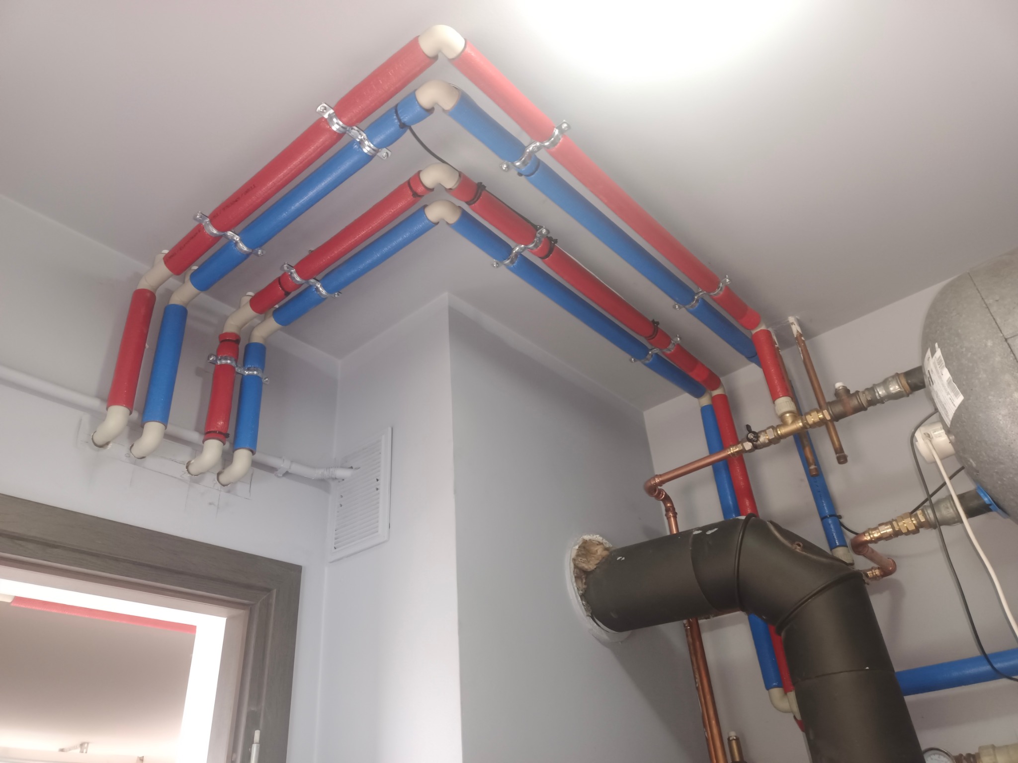 Instalacja grzewcza z rurami w izolacji (czerwone i niebieskie) zamontowanymi pod sufitem, z widocznymi elementami kotła i zaworami. Białe ściany i drzwi w tle.