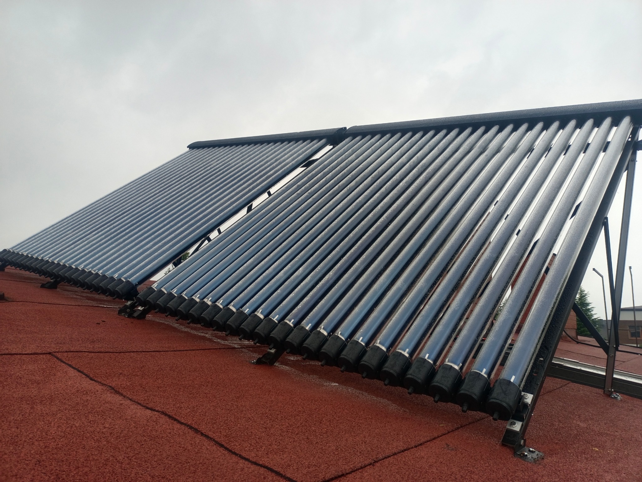 Ustawione na czerwonym dachu, dwa panele kolektorów słonecznych z rurami próżniowymi, lekko pochylone pod kątem, pod pochmurnym niebem.