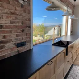 Czarny blat kuchenny z konglomeratu na tle ściany z cegły i widoku na zewnątrz, złota bateria przy czarnym zlewozmywaku, oświetlenie trzema białymi lampami wiszącymi.