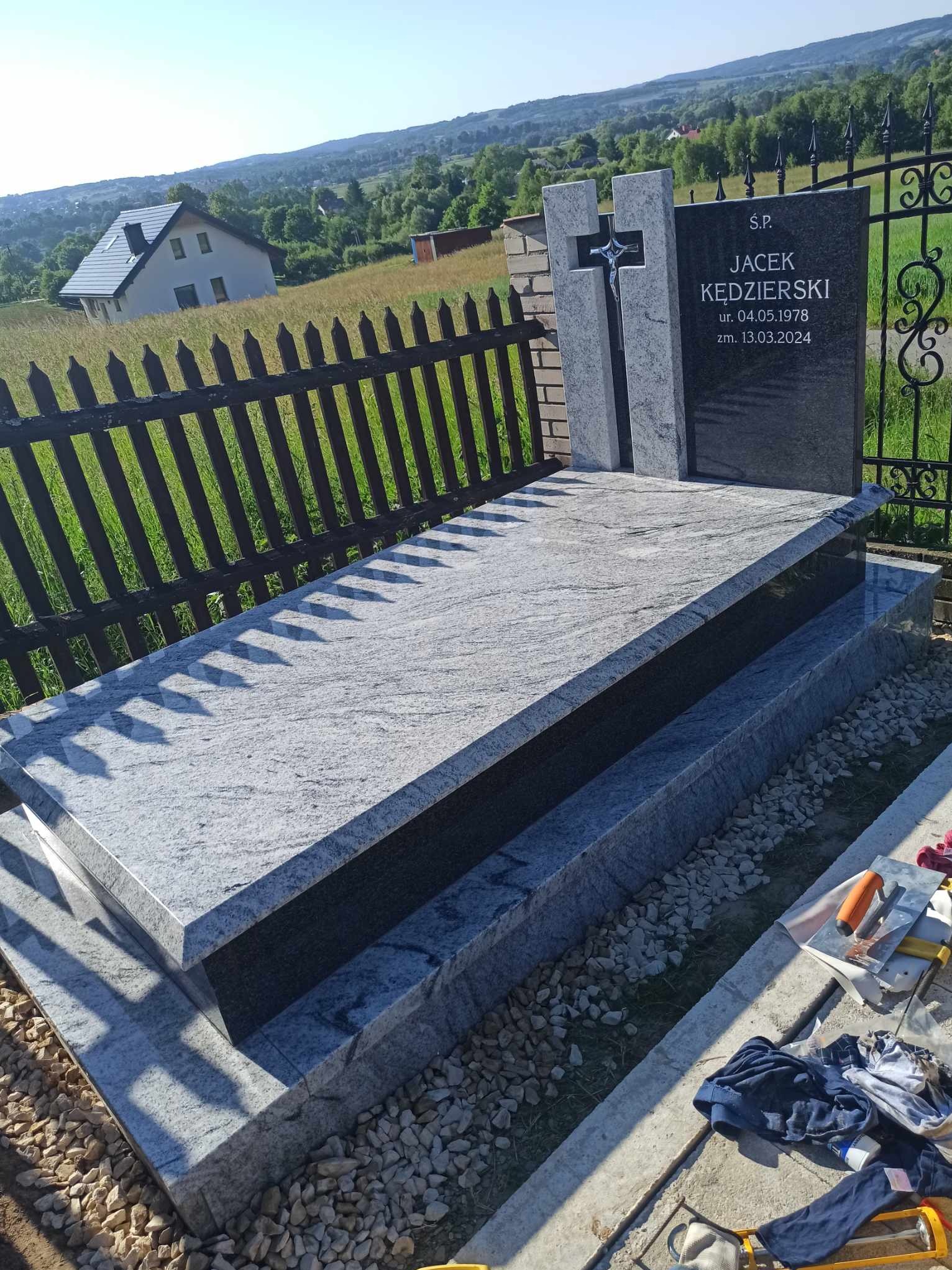 Granitowy pomnik z krzyżem i czarną tablicą nagrobną z inskrypcją, na tle krajobrazu wiejskiego z domem i ogrodzeniem, w pobliżu narzędzia kamieniarskie.