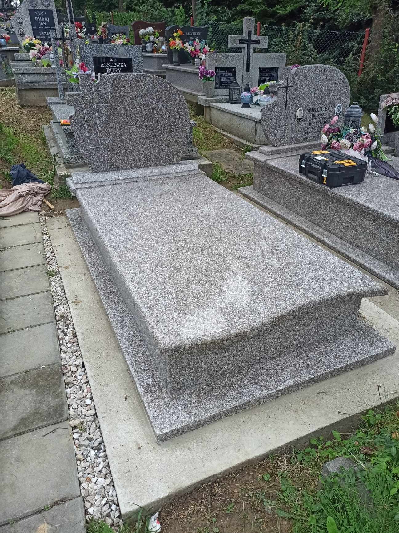 Jasnoszary, granitowy pomnik z krzyżem, otoczony innymi nagrobkami na cmentarzu.