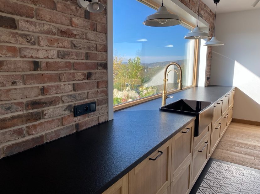 Czarny blat kuchenny z konglomeratu na tle ściany z cegły i widoku na zewnątrz, złota bateria przy czarnym zlewozmywaku, oświetlenie trzema białymi lampami wiszącymi.