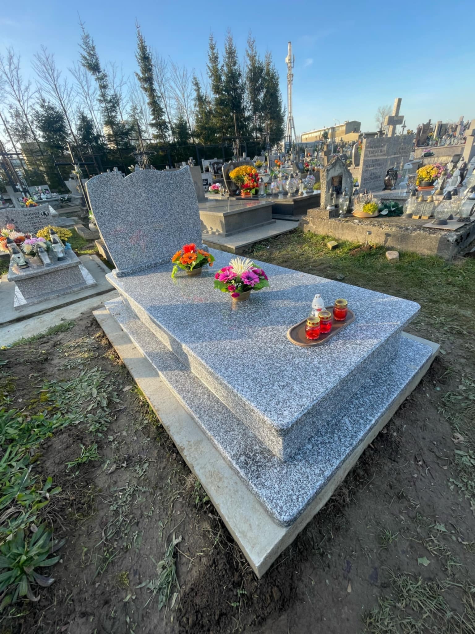 Szary, dwupoziomowy pomnik z granitu na cmentarzu, ozdobiony kwiatami w doniczkach i lampionami na tacy.