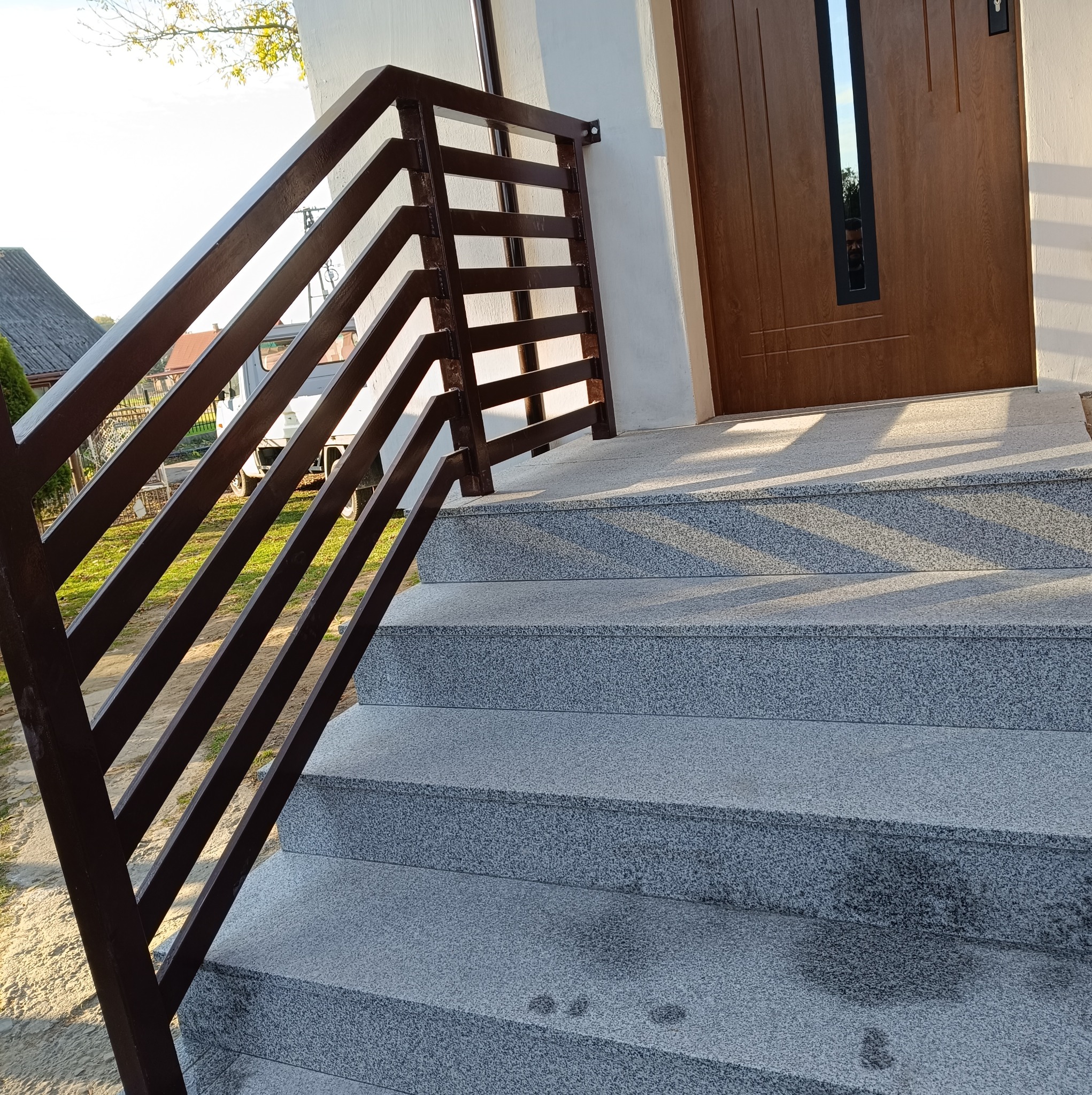 Zewnętrzne schody wejściowe z szarego kamienia z metalową, brązową balustradą, prowadzące do drewnianych drzwi wejściowych z czarnym, pionowym panelem okiennym.