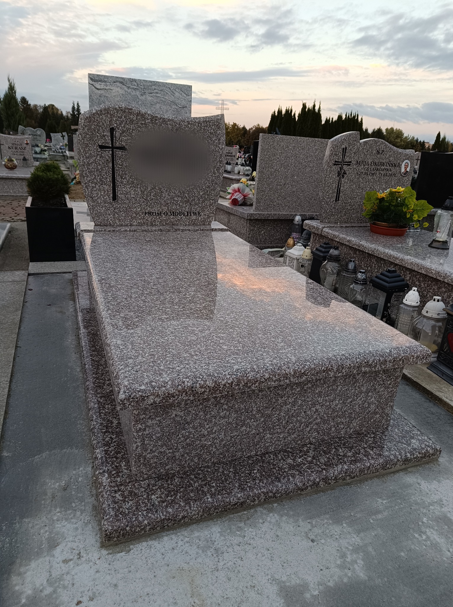 Polerowany, dwupoziomowy pomnik z szarego granitu z krzyżem, widoczny na tle cmentarza o zmierzchu.