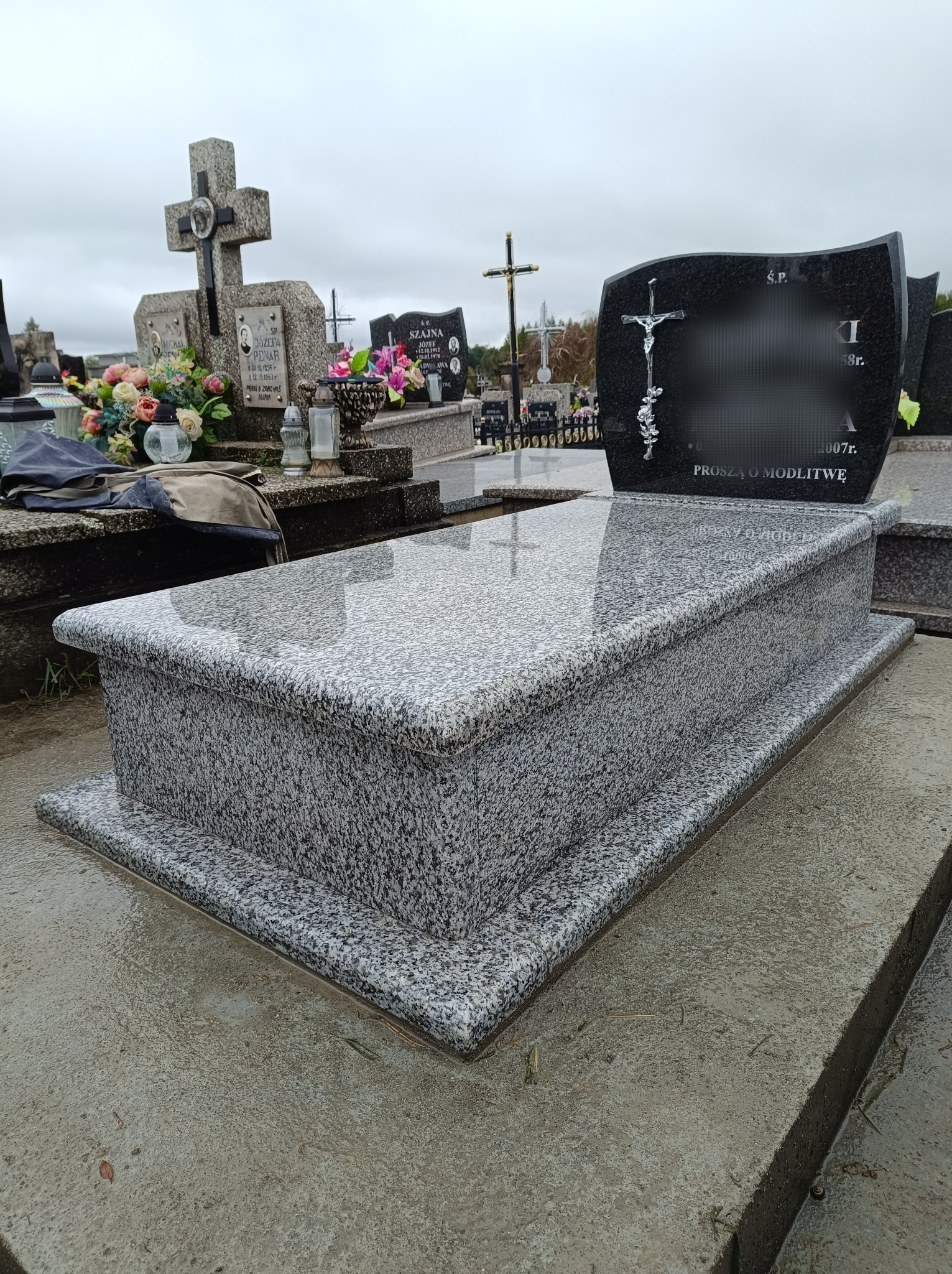 Granitowy grobowiec z ciemnoszarą płytą nagrobkową, ozdobioną srebrnym krucyfiksem i napisem 'Proszą o modlitwę', na cmentarzu w pochmurny dzień.