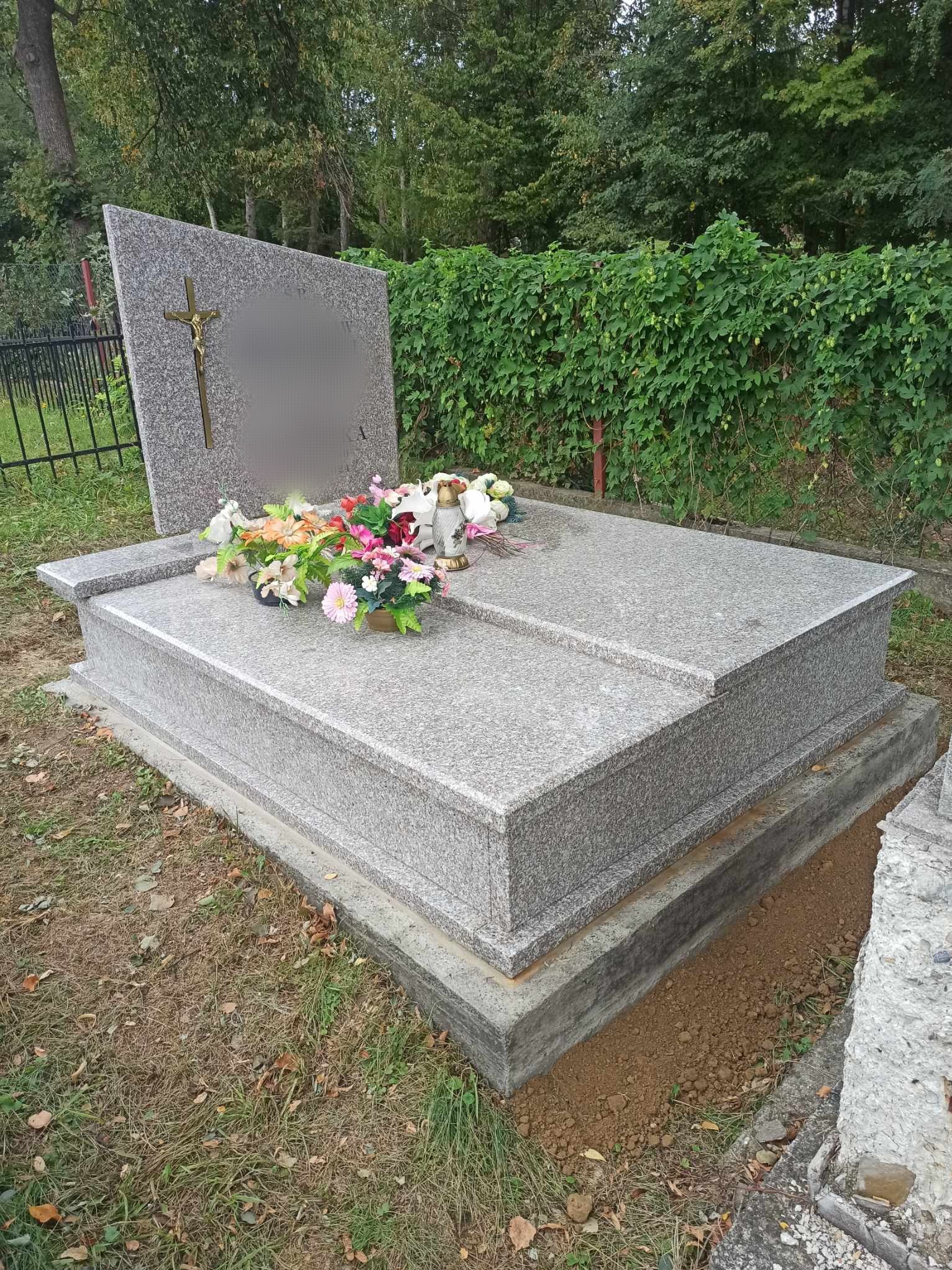 Granitowy nagrobek z krzyżem i kwiatami, otoczony zielenią, widok z boku.