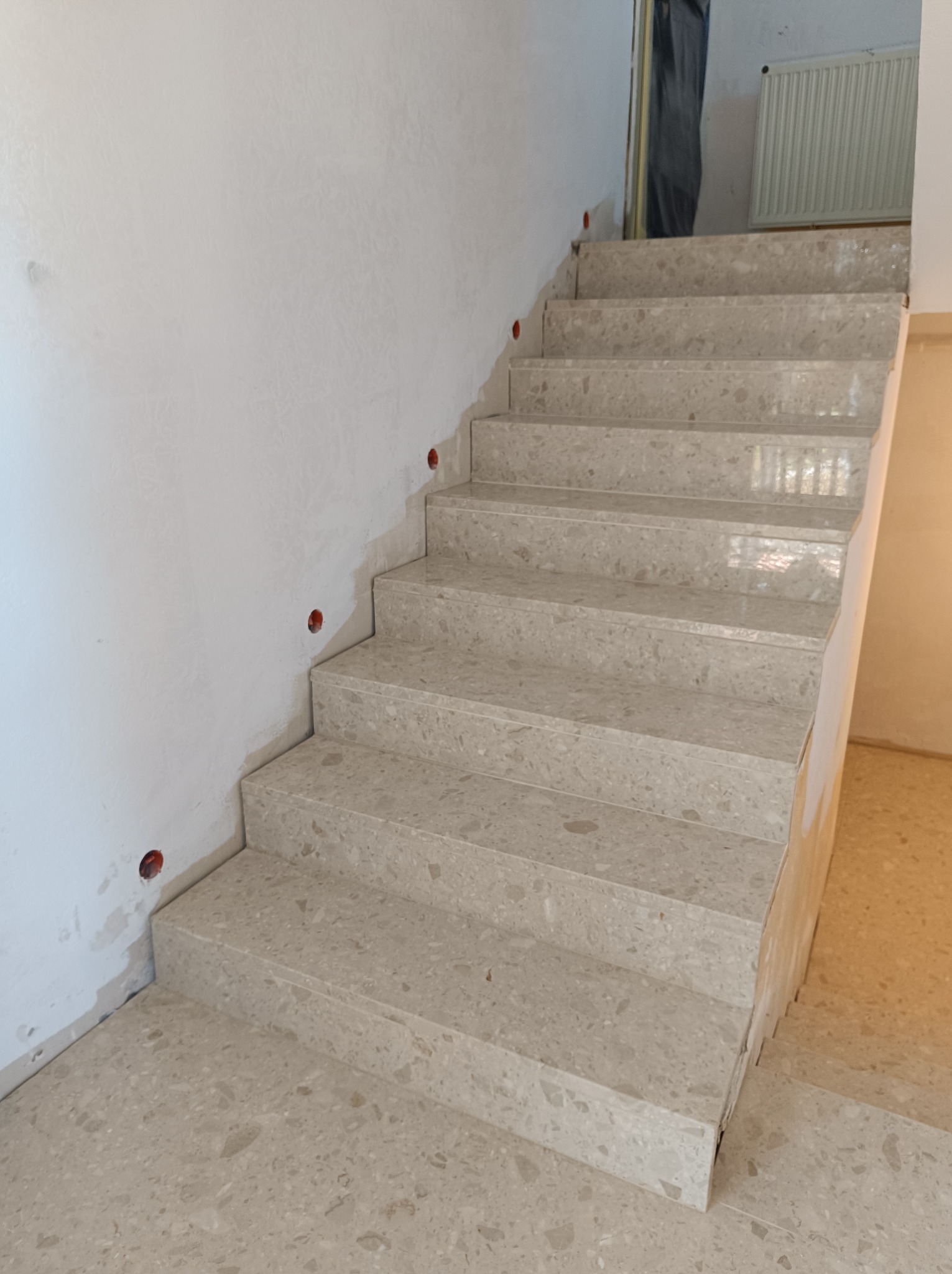 Wewnętrzne schody z jasnego kamienia kompozytowego o drobnej fakturze, widoczne otwory na puszki elektryczne w ścianie, etap wykończeniowy.
