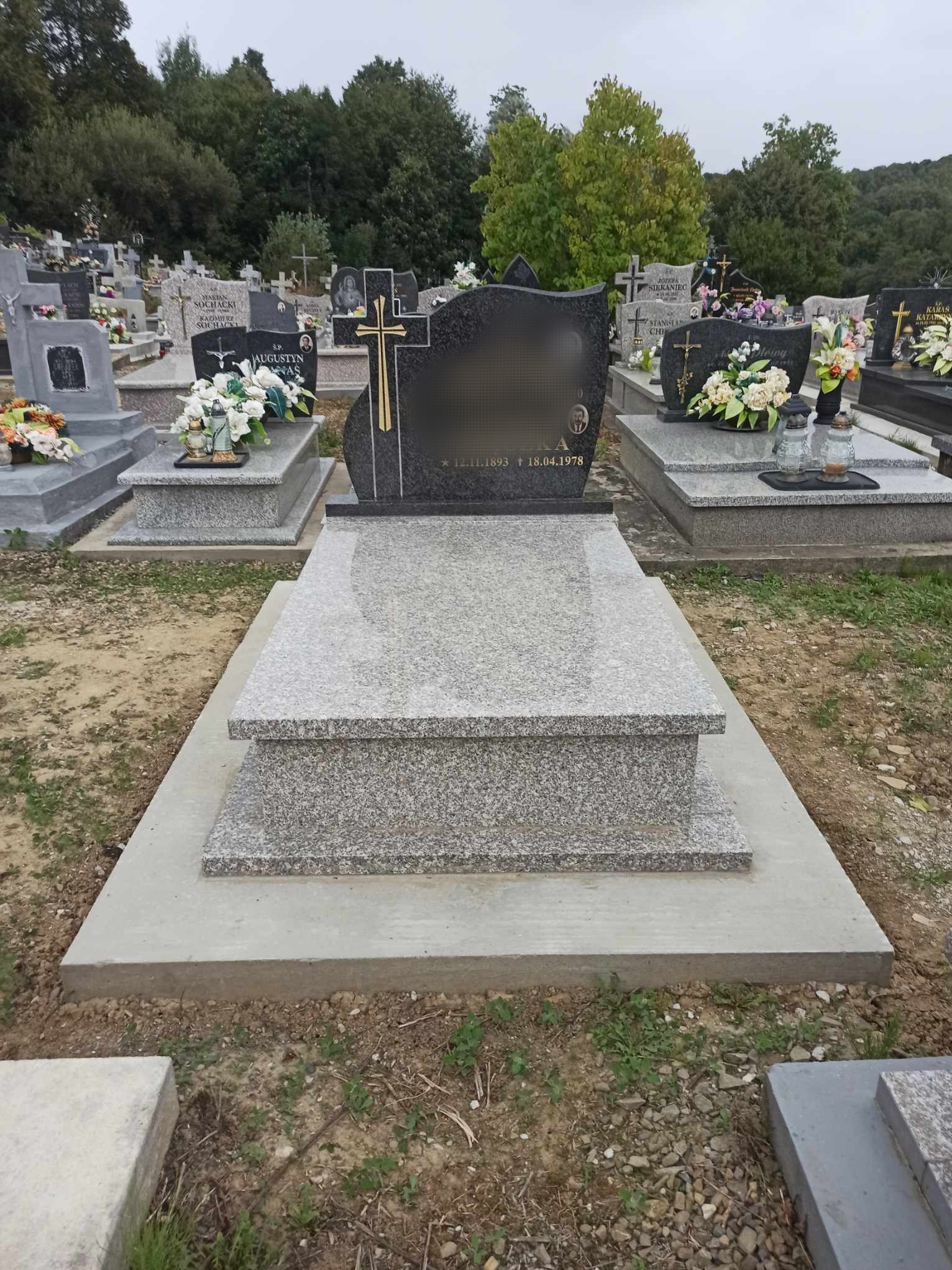 Granitowy nagrobek z czarną tablicą i złotym krzyżem na cmentarzu, widoczne inskrypcje i inne nagrobki w tle.