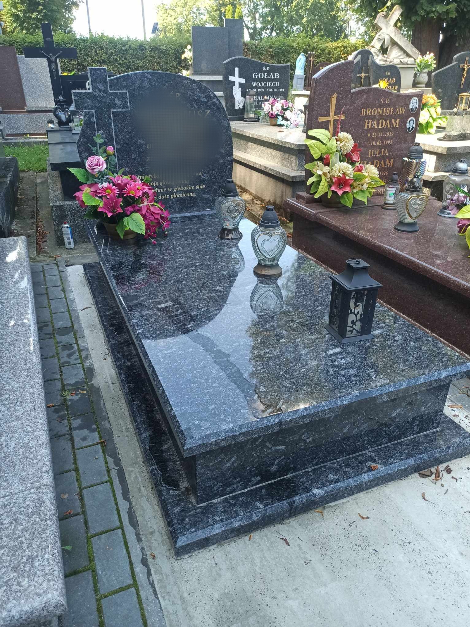 Granitowy nagrobek z ciemnego kamienia z kwiatami w doniczce i lampionem, odbijający światło, na tle innych nagrobków na cmentarzu.