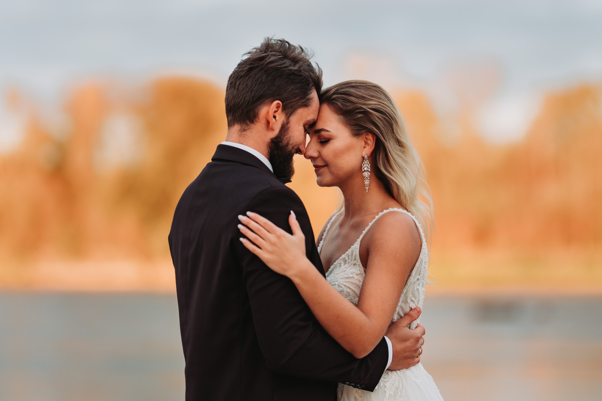 Para młoda, on w czarnym garniturze, ona w białej sukni z koronkowymi zdobieniami i długimi kolczykami, obejmują się czule czołami w plenerze z rozmytym tłem w odcieniach pomarańczy i błękitu.