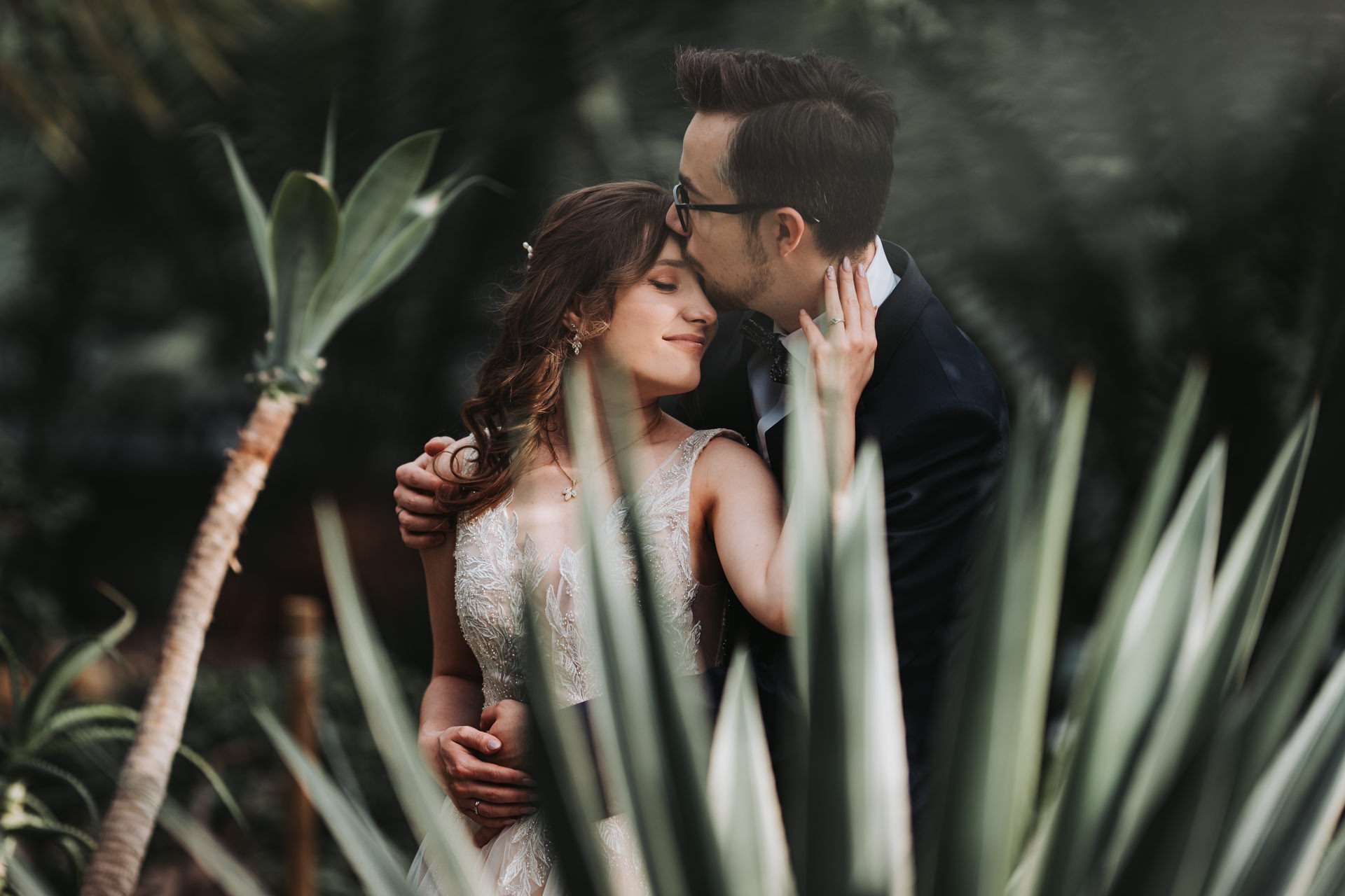 Para młoda w objęciach w otoczeniu egzotycznej roślinności: on w granatowym garniturze i okularach całuje ją w czoło, ona w zdobnej sukni ślubnej z zamkniętymi oczami.
