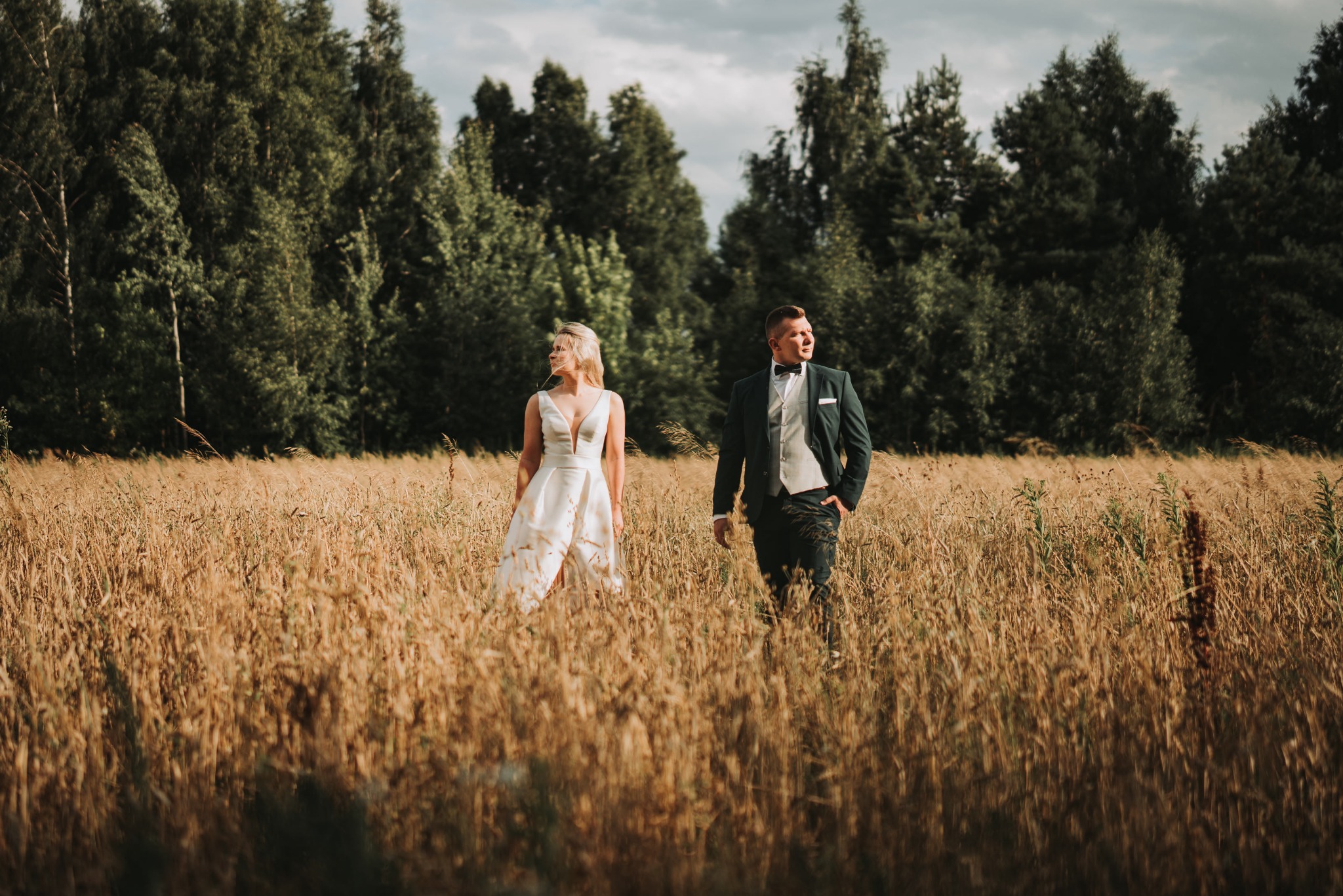 Para młoda spaceruje po polu zboża w słoneczny dzień, drzewa w tle, on w ciemnozielonym garniturze, ona w białej sukni z głębokim dekoltem.