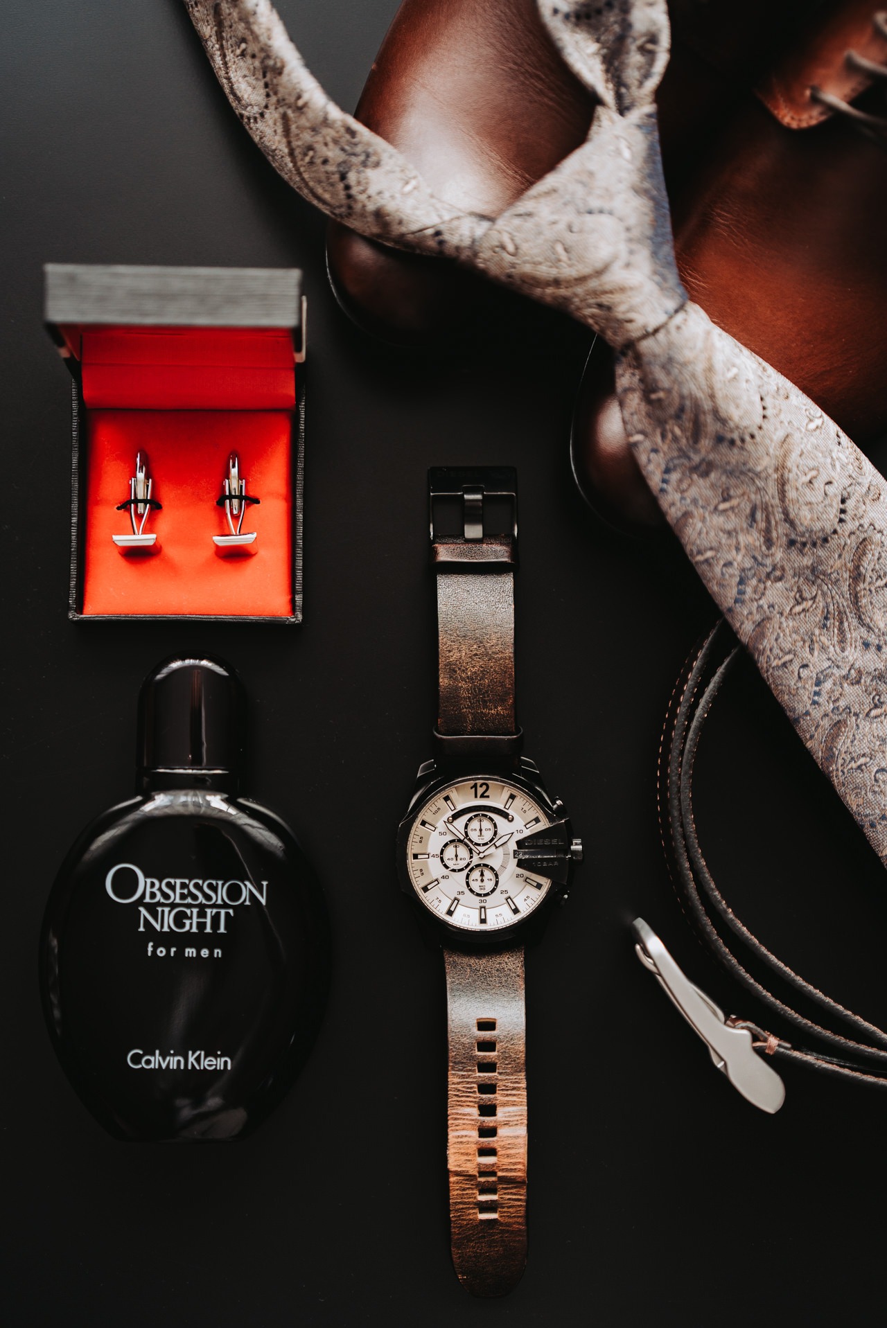 Męskie akcesoria na czarnym tle: skórzane buty, wzorzysty krawat, spinki do mankietów w pudełku, zegarek Diesel z brązowym paskiem, pasek i flakon perfum Calvin Klein Obsession Night.