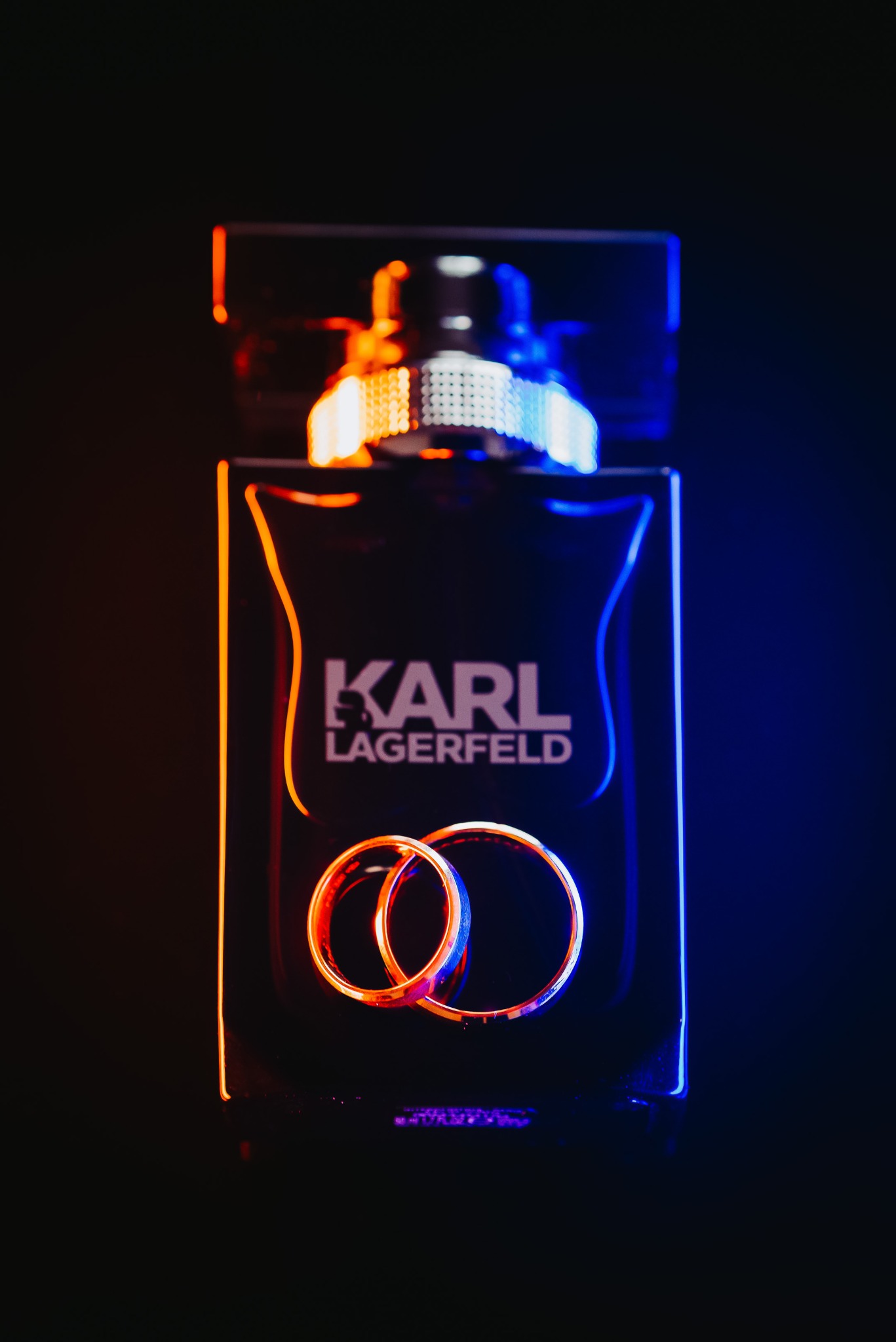 Dwa złote obrączki ślubne opierają się o flakon perfum Karl Lagerfeld na czarnym tle, oświetlone pomarańczowym i niebieskim światłem.