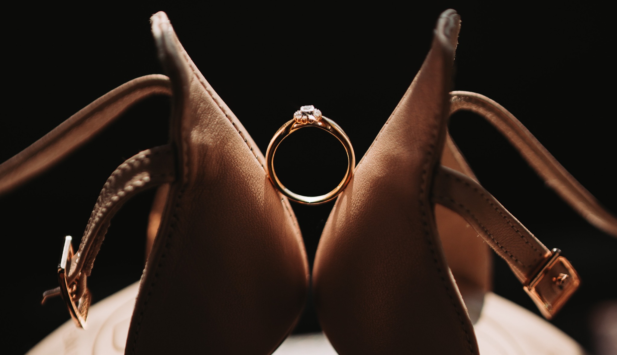 Złoty pierścionek zaręczynowy z brylantem spoczywa między czubkami beżowych, skórzanych butów na obcasie, zapiętych na kostce, na ciemnym tle.