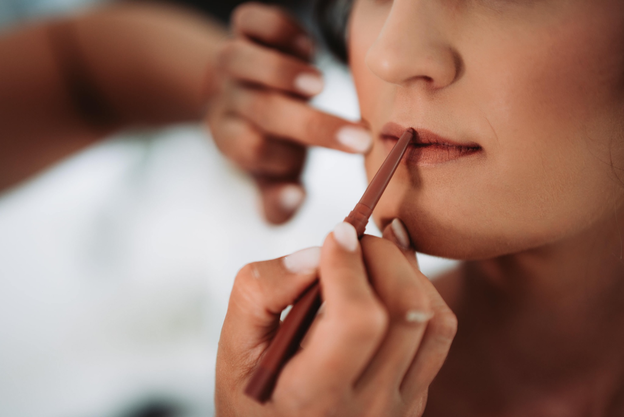 Makijażystka podkreśla usta klientki konturówką w odcieniu nude, zbliżenie na twarz i dłonie, widoczny pierścionek na palcu