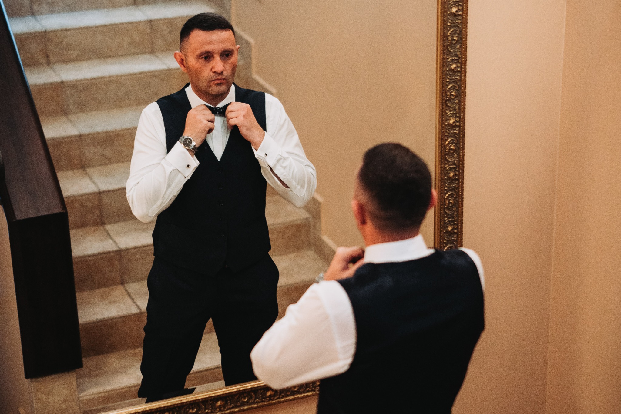 Mężczyzna w czarnej kamizelce i białej koszuli, z zegarkiem na nadgarstku, poprawia muchę przed lustrem w złotej ramie, na tle schodów.