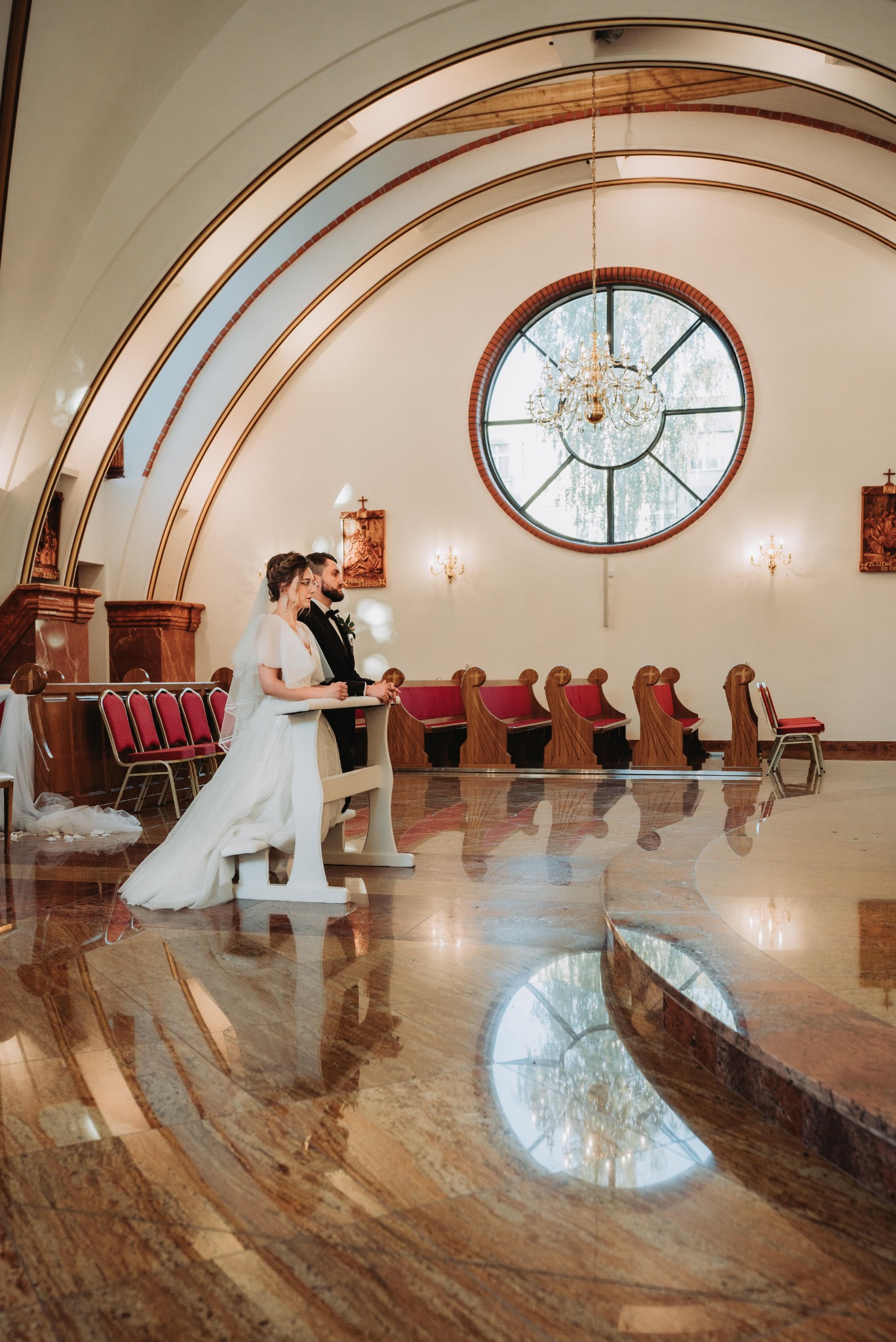 Para młoda w kościele podczas ceremonii, panna młoda w sukni z bufiastymi rękawami i welonem, pan młody w czarnym garniturze, marmurowa posadzka odbija światło, w tle okrągłe okno z żyrandolem.