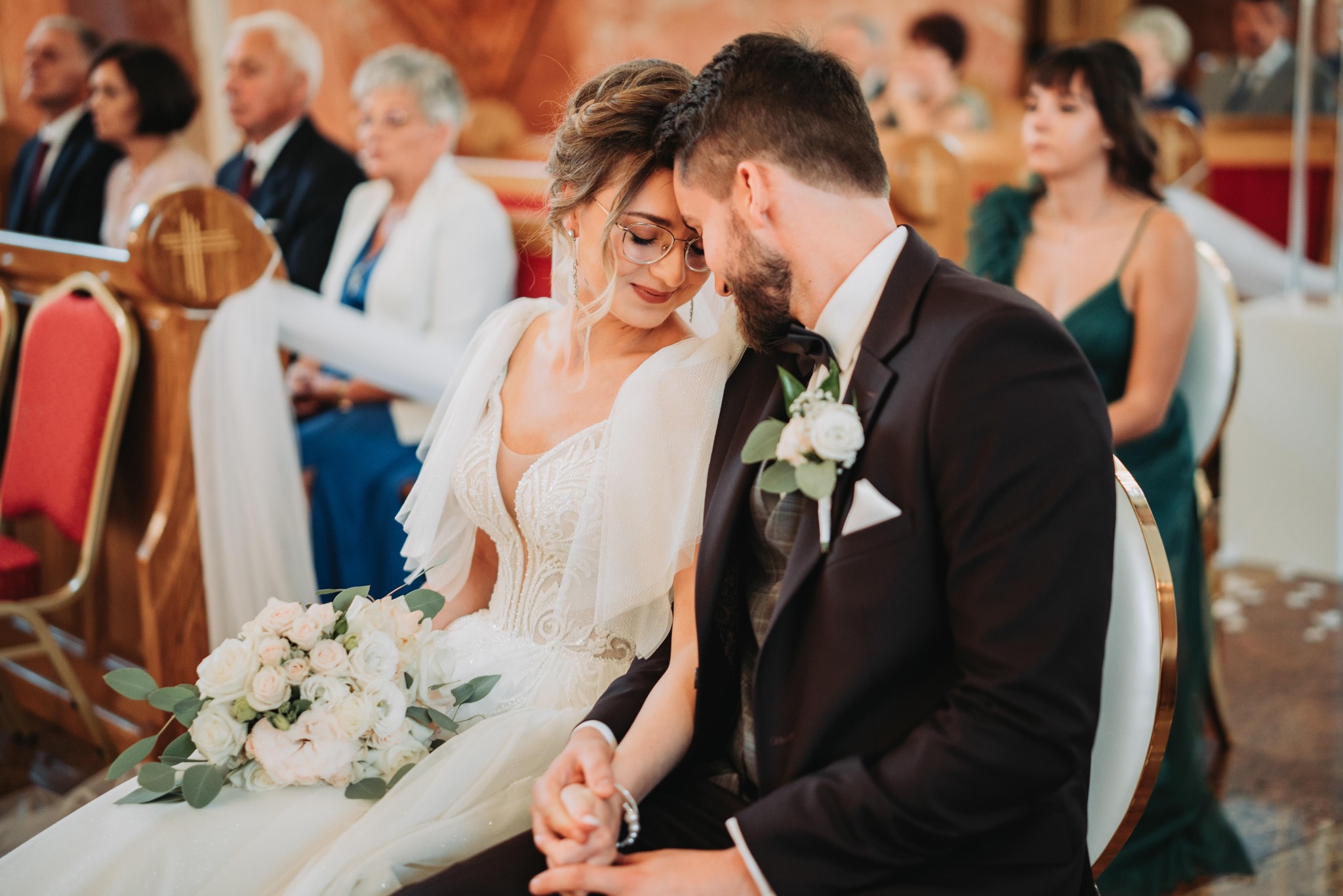 Uczucie i bliskość nowożeńców w kościele w Mińsku Mazowieckim. Panna młoda w okularach i bogato zdobionej sukni trzyma bukiet, pan młody w ciemnym garniturze z butonierką. W tle goście siedzący...