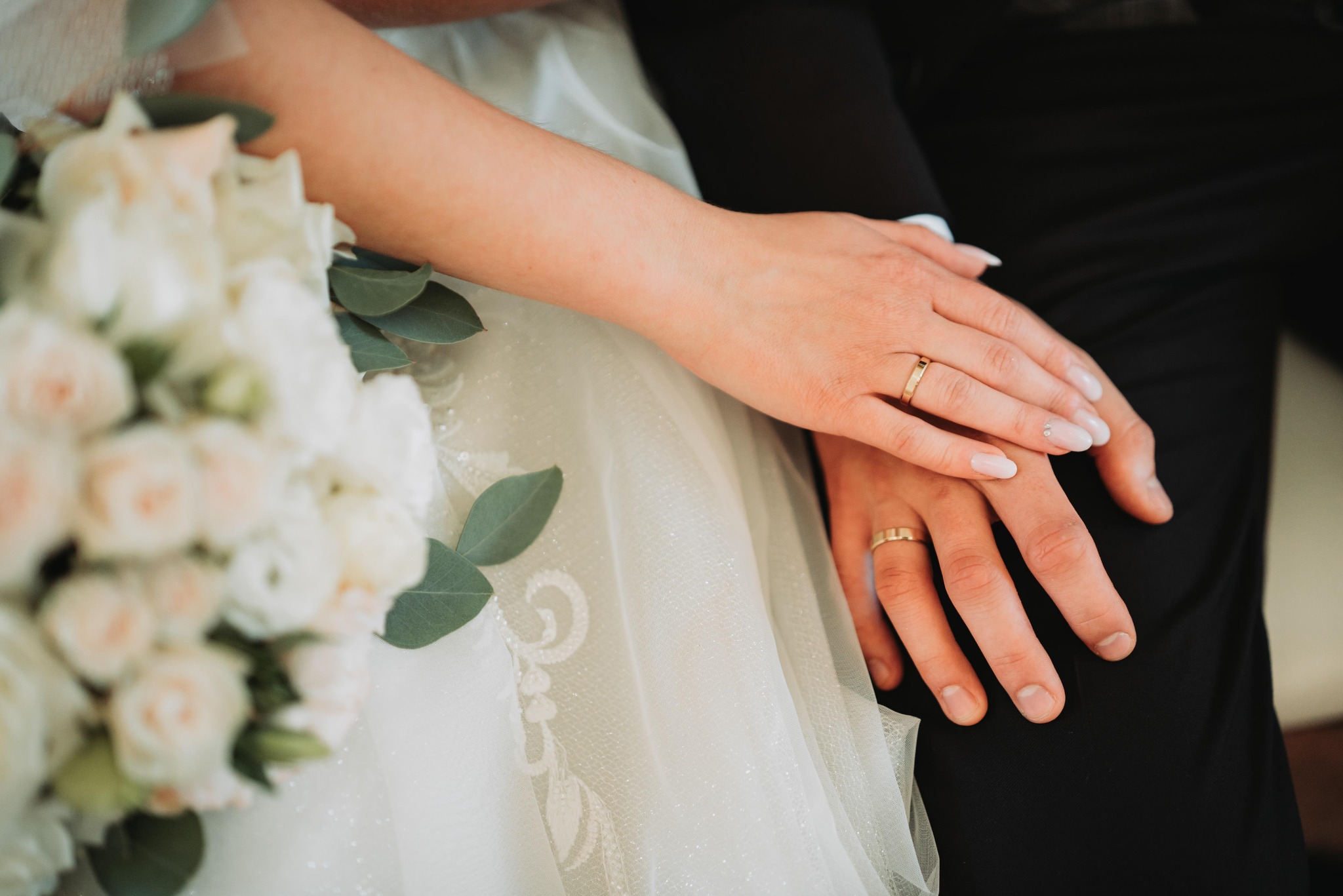 Dłonie pary młodej z obrączkami na tle bukietu ślubnego i sukni z delikatną koronką.