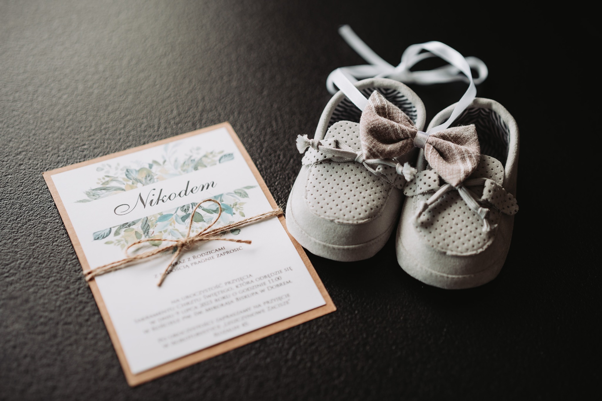 Zaproszenie na chrzest o imieniu Nikodem, przewiązane sznurkiem, leżące obok pary dziecięcych bucików z kokardkami na ciemnym tle.