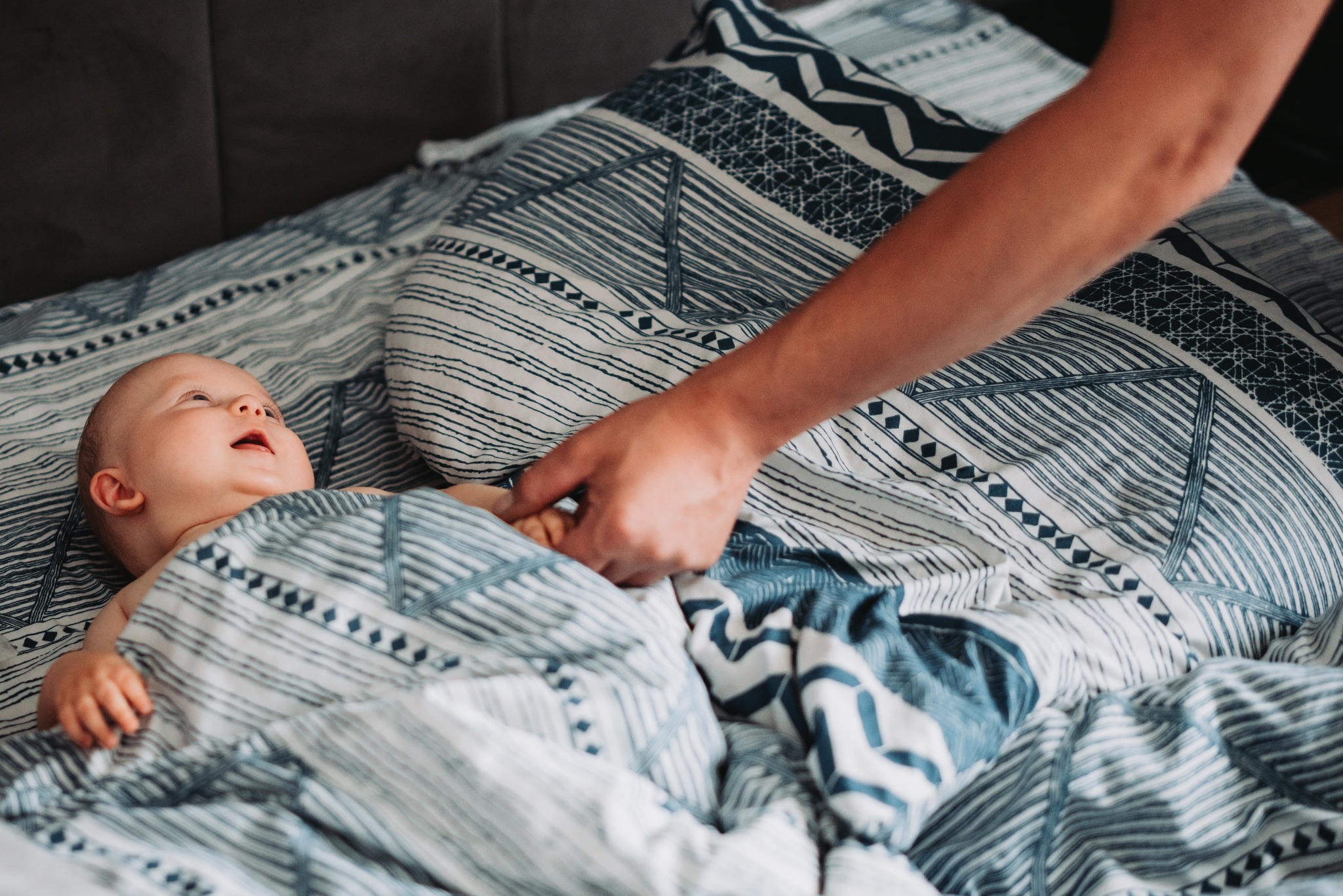 Uśmiechnięte niemowlę leżące na wzorzystej pościeli, trzymane za rękę przez dorosłego