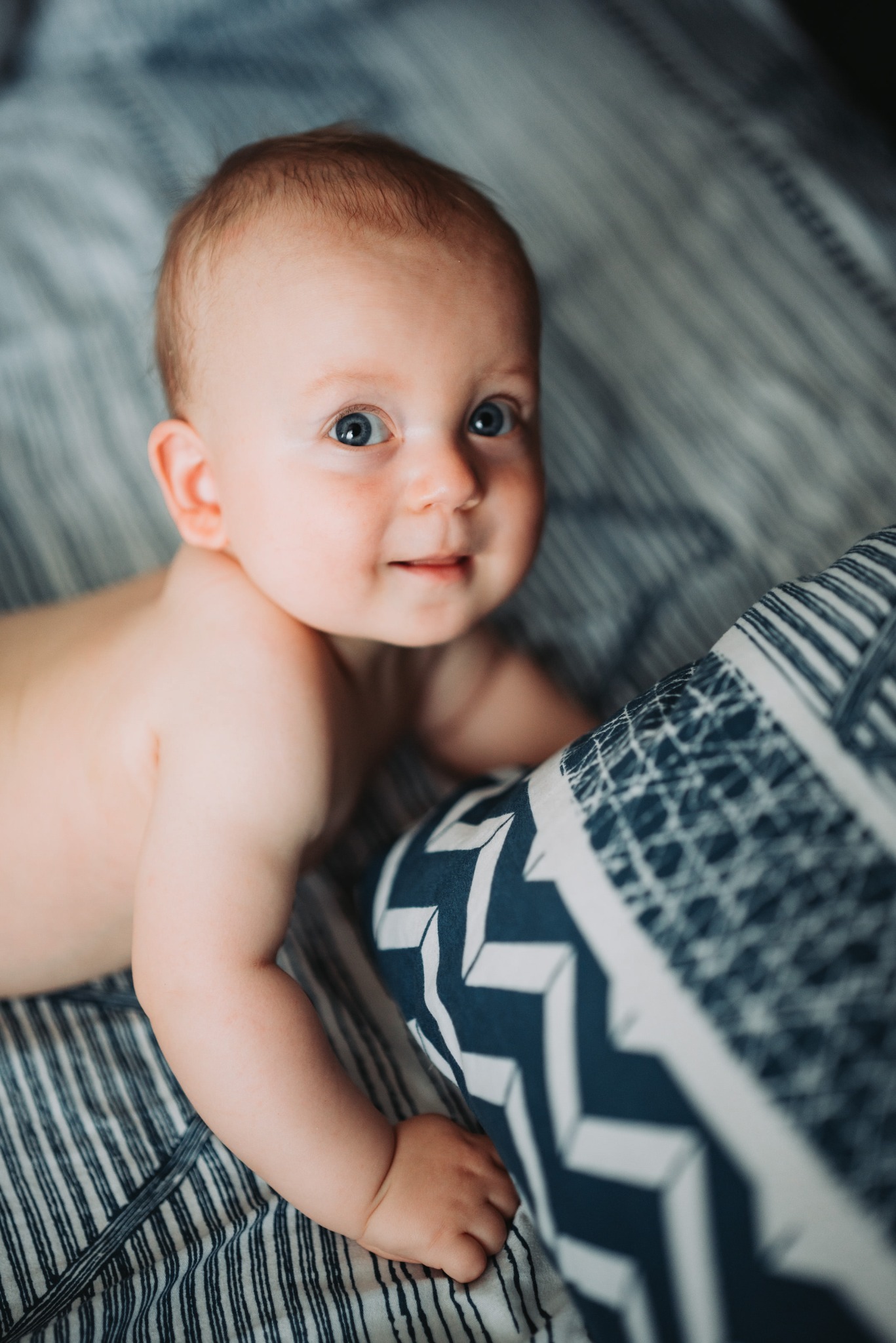 Uśmiechnięty niemowlak z niebieskimi oczami leży na boku na pościeli w paski, opierając się o poduszkę w geometryczne wzory.