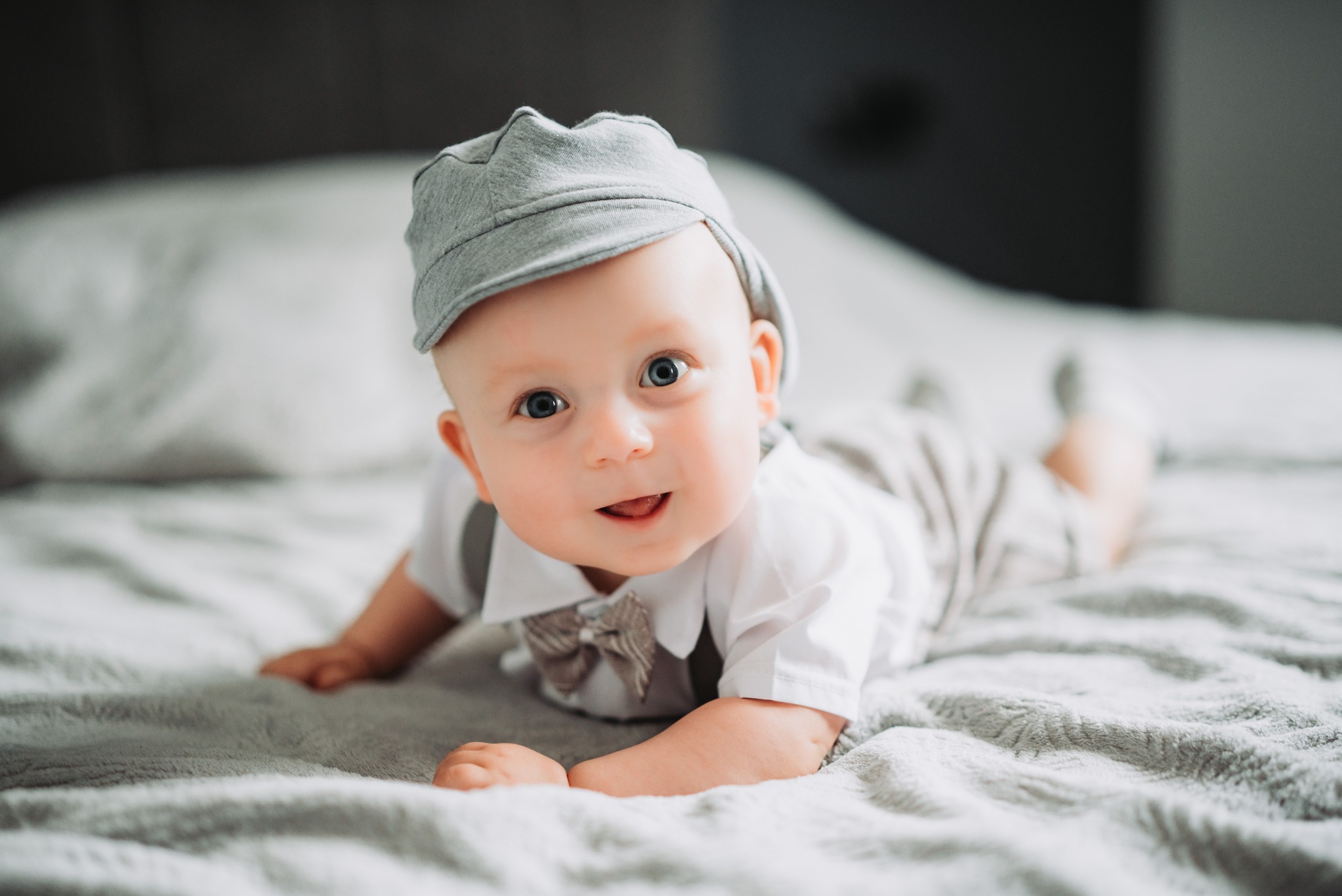 Uroczy niemowlak w szarym kapeluszu i muszce leżący na brzuchu na szarym kocu, z otwartymi ustami i patrzący prosto w obiektyw.
