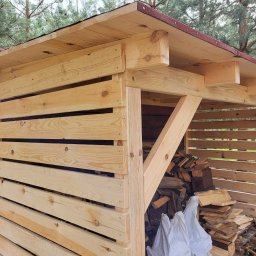 Usługi ogólnobudowlane Artur Klecha - Drewniana konstrukcja z dachem krytym papą, prawdopodobnie wiata na drewno, wypełniona pociętymi kawałkami drewna i zabezpieczona białym workiem, widoczna na tle drzew.