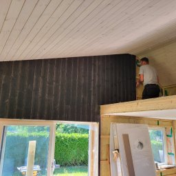 Usługi ogólnobudowlane Artur Klecha - Wnętrze domu w trakcie budowy, widoczna antresola z drewnianą konstrukcją, ściany wykończone ciemnym drewnem, biały sufit z drewnianych paneli, okno z widokiem na zieleń, drabina i materiały...